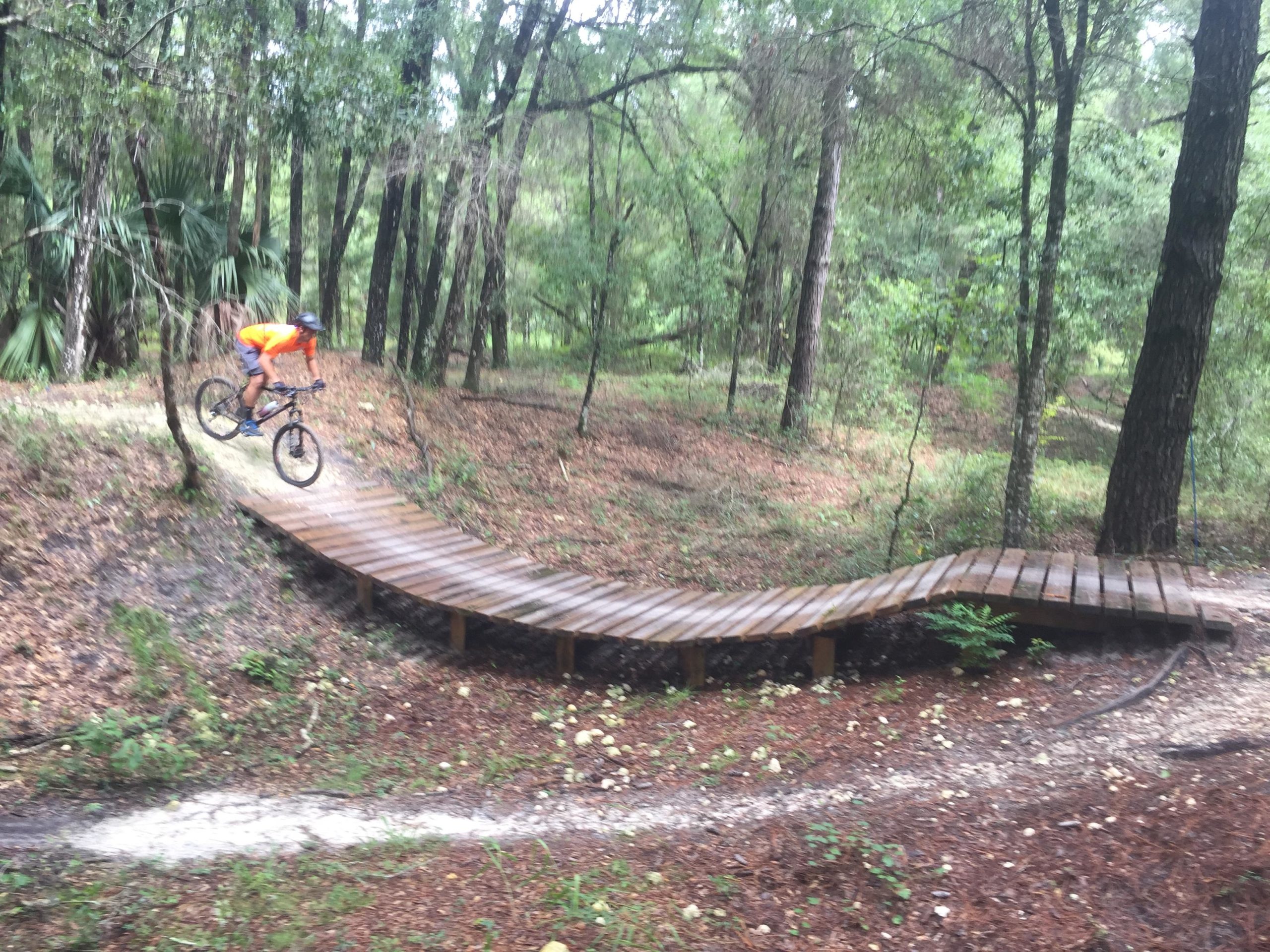 A person in an orange shirt and black helmet jumping off a wooden ramp while riding a mountain bike in a forested area. The ground is covered in leaves and the scene features lush green trees in the background. Santos mountain bike trail.