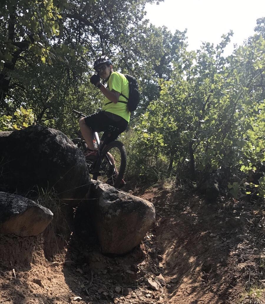 A person wearing a bright yellow shirt and a helmet is sitting on a mountain bike, positioned on a large rock in a natural outdoor setting. They are giving a thumbs-up gesture while surrounded by trees and uneven terrain. The sunlight is shining through the foliage, creating a lively atmosphere. Lake Lawtonka Trails mountain bike trail.