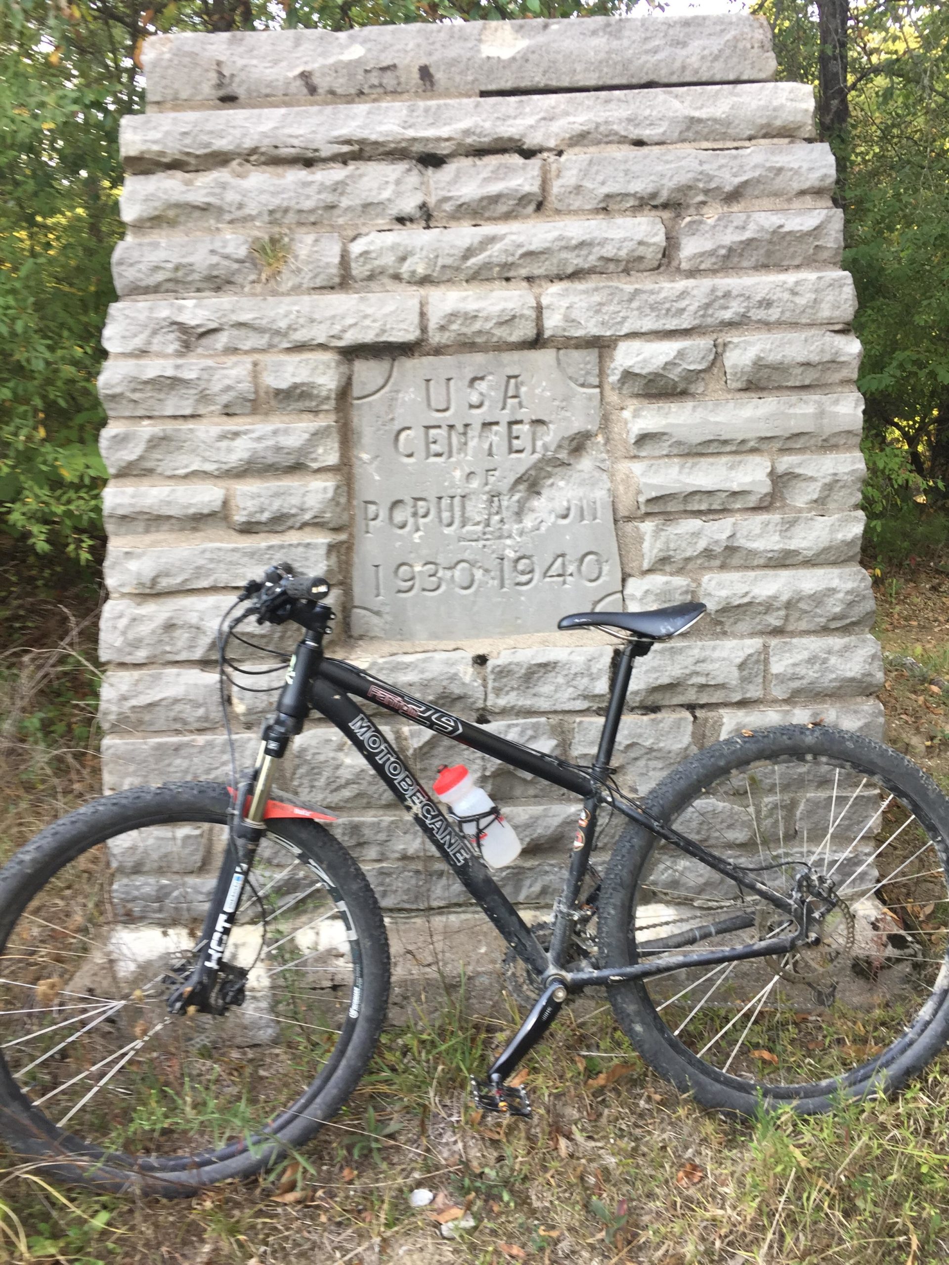 A mountain bike leans against a stone monument with the inscription "USA Center of Population 1930-1940," surrounded by greenery and grass. Linton Conservation Club mountain bike trail.