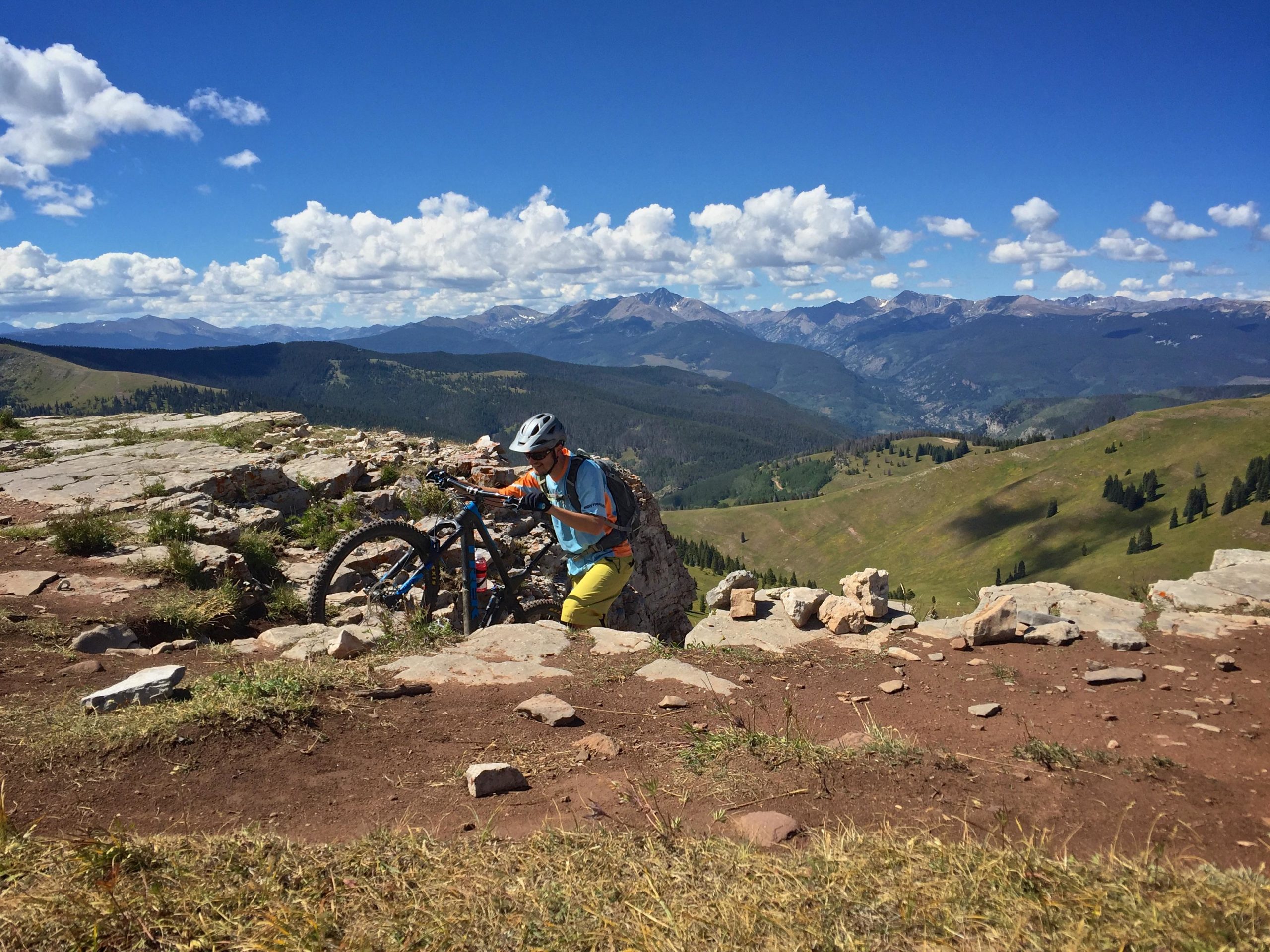 A mountain biker in a bright blue shirt and yellow pants is adjusting their bike while standing on rocky terrain, with a scenic backdrop of lush green hills, distant mountains, and a vibrant blue sky dotted with fluffy white clouds. Two Elk via Vail Pass mountain bike trail.