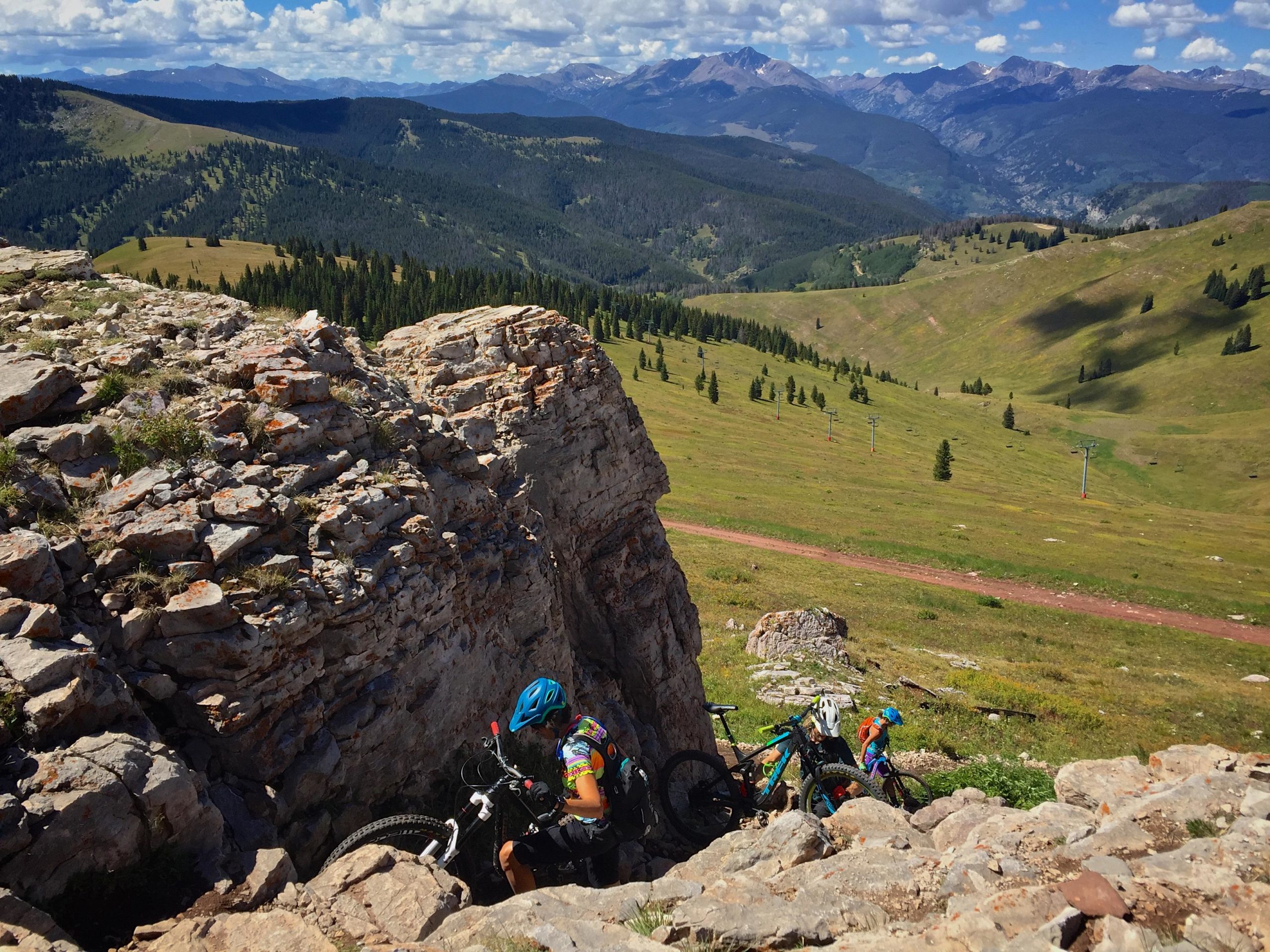 A mountain biker navigates a rocky outcrop while two bicycles rest nearby, set against a backdrop of expansive green hills and distant mountains under a partly cloudy sky. Two Elk via Vail Pass mountain bike trail.