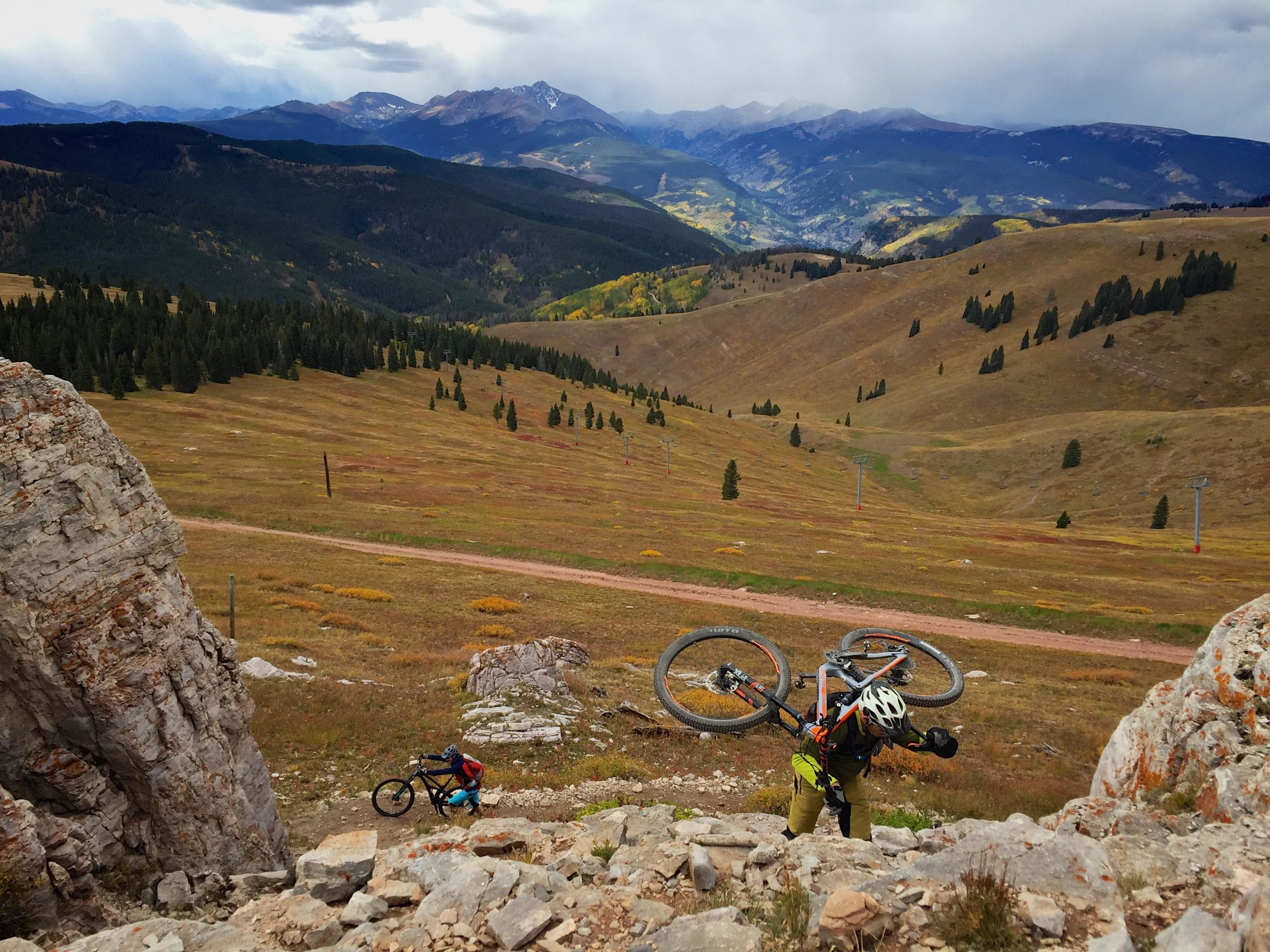 Two mountain bikers navigate a rocky ascent in a mountainous landscape, carrying their bikes. The background features rolling hills covered in greenery and patches of golden foliage, with distant mountains under a cloudy sky. Vail Mountain Bike Park mountain bike trail.