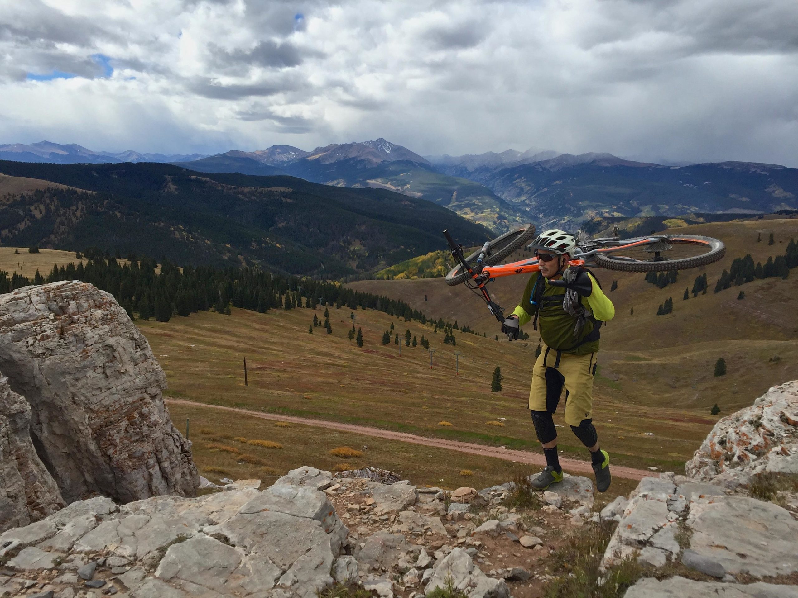 A mountain biker carrying a bike over rocky terrain, with a scenic view of mountains and valleys in the background, set against a cloudy sky. Vail Mountain Bike Park mountain bike trail.