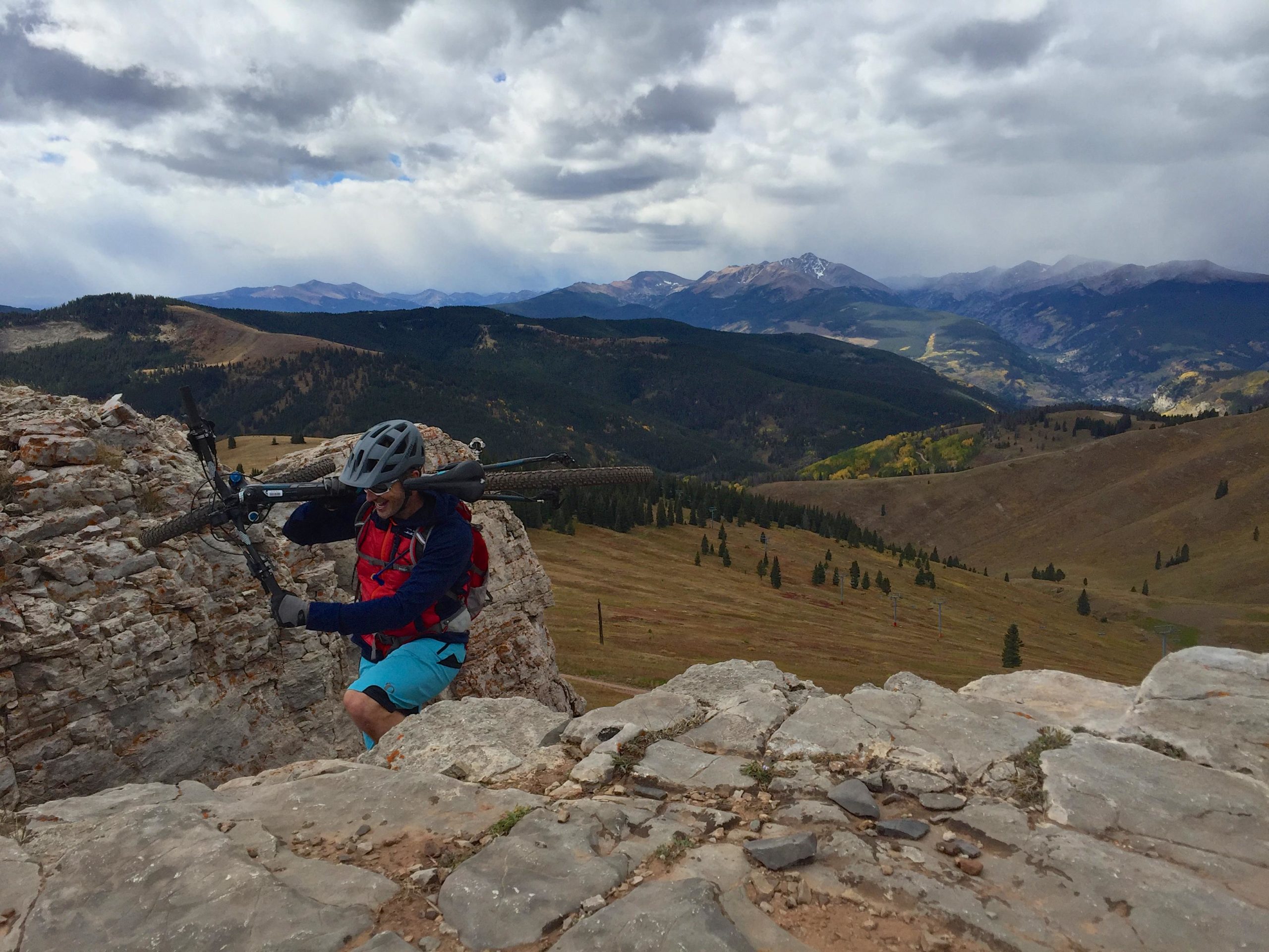 A mountain biker navigating rocky terrain while carrying a bicycle on his shoulder, with a panoramic view of mountains and valleys in the background under a partly cloudy sky. Vail Mountain Bike Park mountain bike trail.