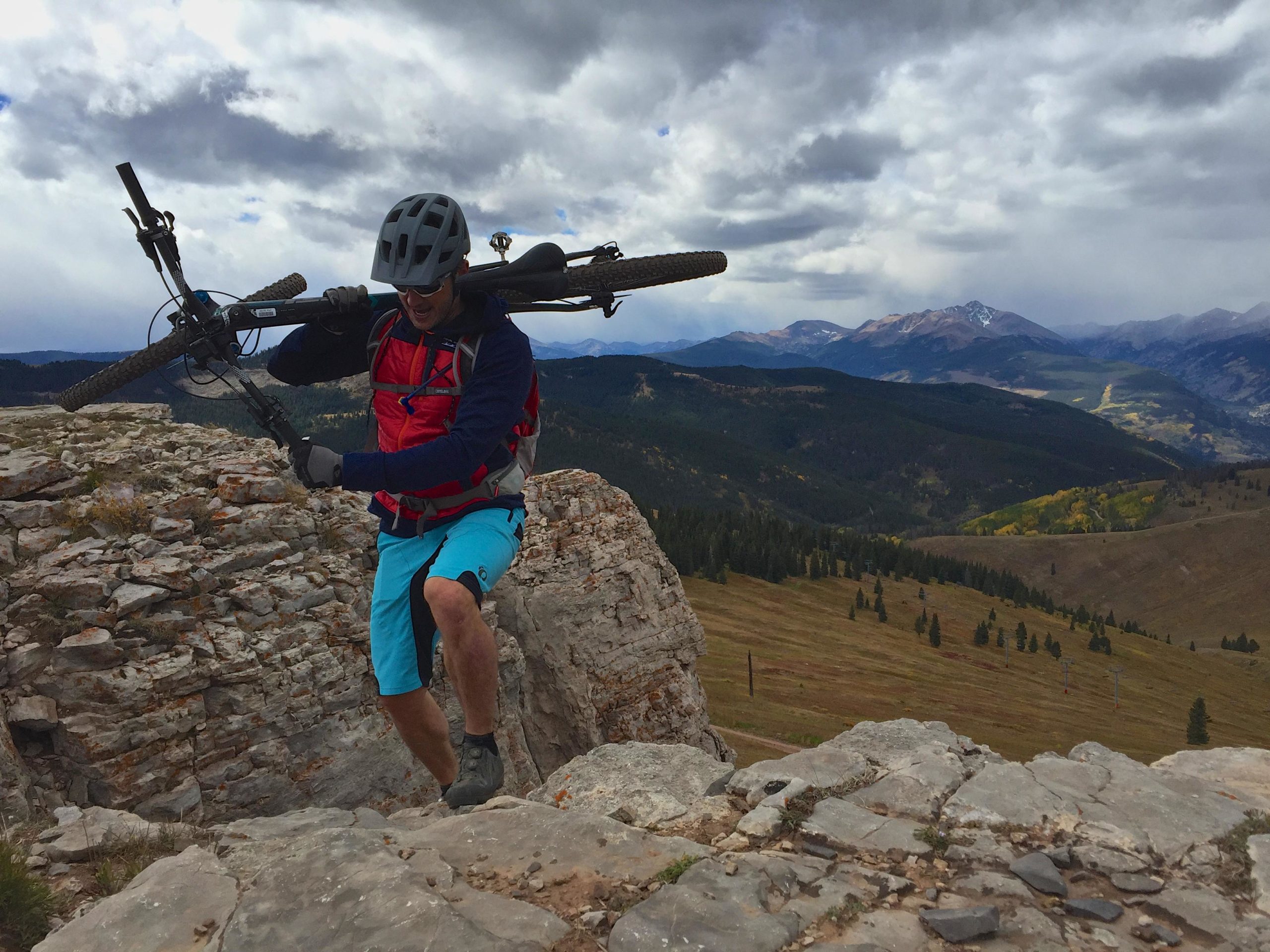 A mountain biker carrying a bike while climbing rocky terrain, with a scenic view of mountains and a cloudy sky in the background. The biker is wearing a helmet, a red vest, and blue shorts, showcasing the adventurous and rugged environment. Vail Mountain Bike Park mountain bike trail.