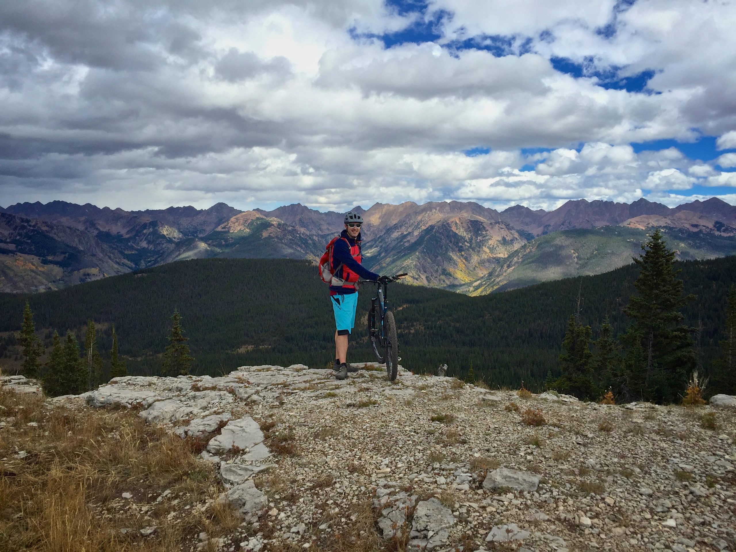 A person stands on a rocky outcrop with a mountain bike, enjoying a scenic view of rugged mountains and a forested valley under a partly cloudy sky. Vail Mountain Bike Park mountain bike trail.