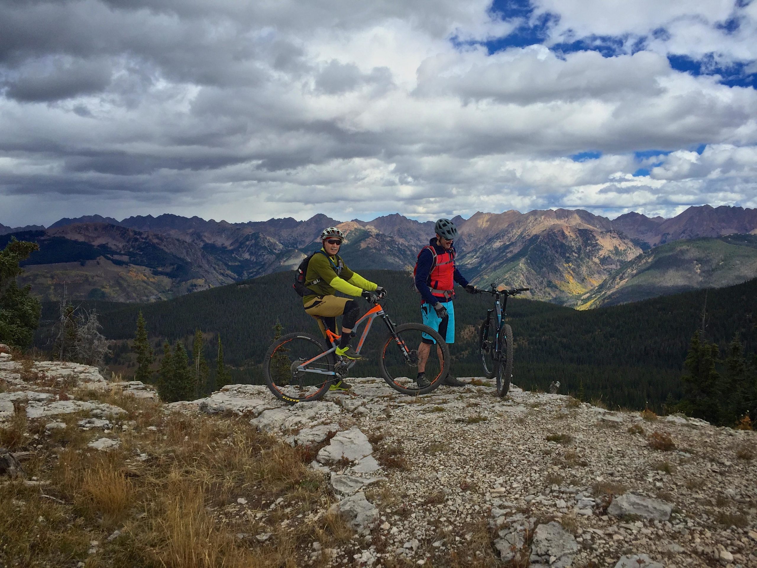 Two mountain bikers stand on a rocky cliff overlooking a mountainous landscape. One rider is wearing a green jacket and brown pants, while the other is dressed in a red vest and blue shorts. The sky is partially cloudy, with darker clouds overhead, and the mountains in the background display a mix of green forests and rocky terrain. Vail Mountain Bike Park mountain bike trail.