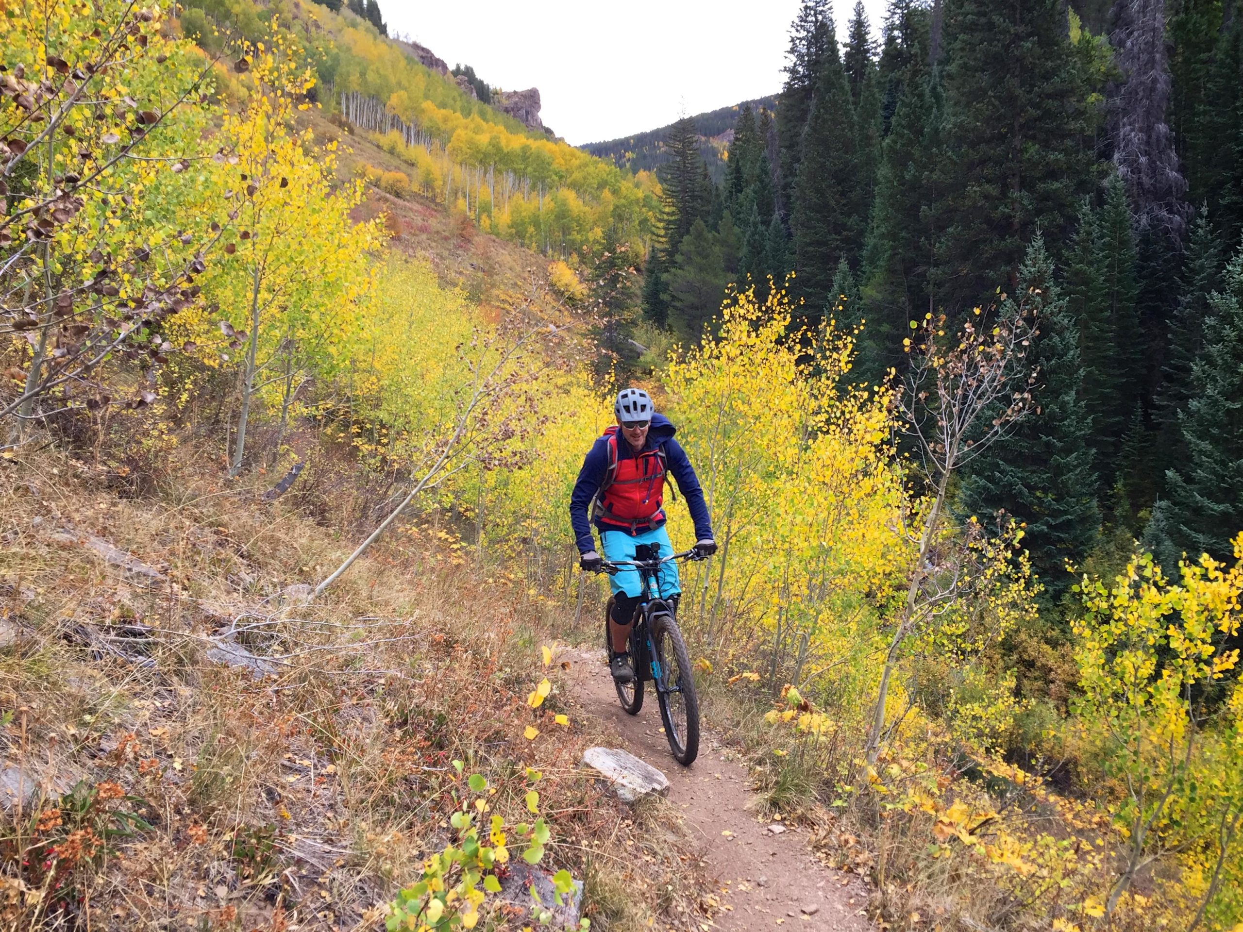 A mountain biker navigating a dirt trail surrounded by vibrant autumn foliage, featuring bright yellow and green trees. The cyclist is wearing a blue helmet, a blue and red outfit, and is focused on the path ahead as he rides through a scenic mountainous landscape. Two Elk via Vail Pass mountain bike trail.