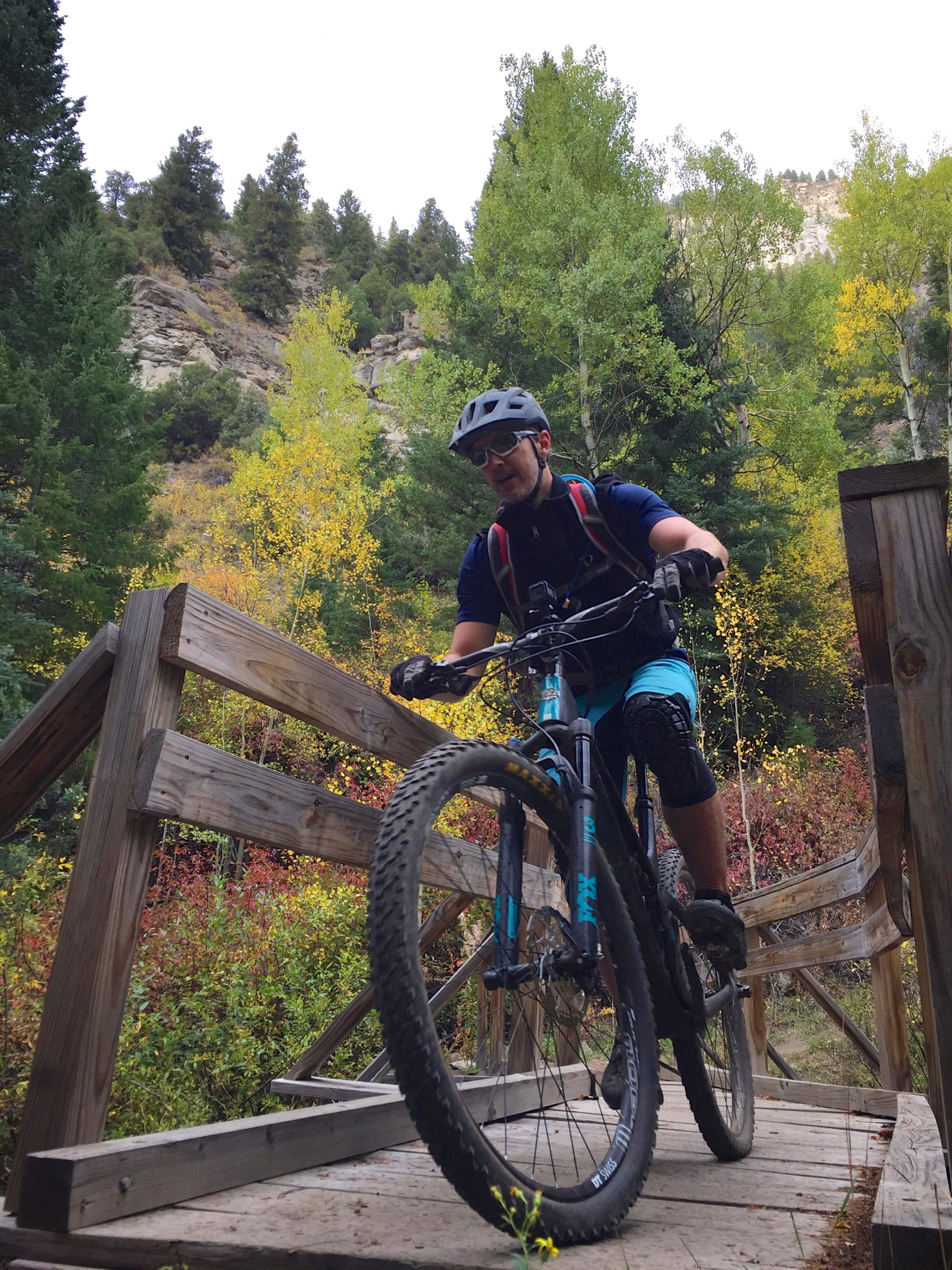 A mountain biker riding over a wooden bridge surrounded by colorful autumn foliage and rocky terrain in a forested area. The cyclist is wearing a helmet, sunglasses, and protective gear, navigating the trail with confidence. Two Elk via Vail Pass mountain bike trail.