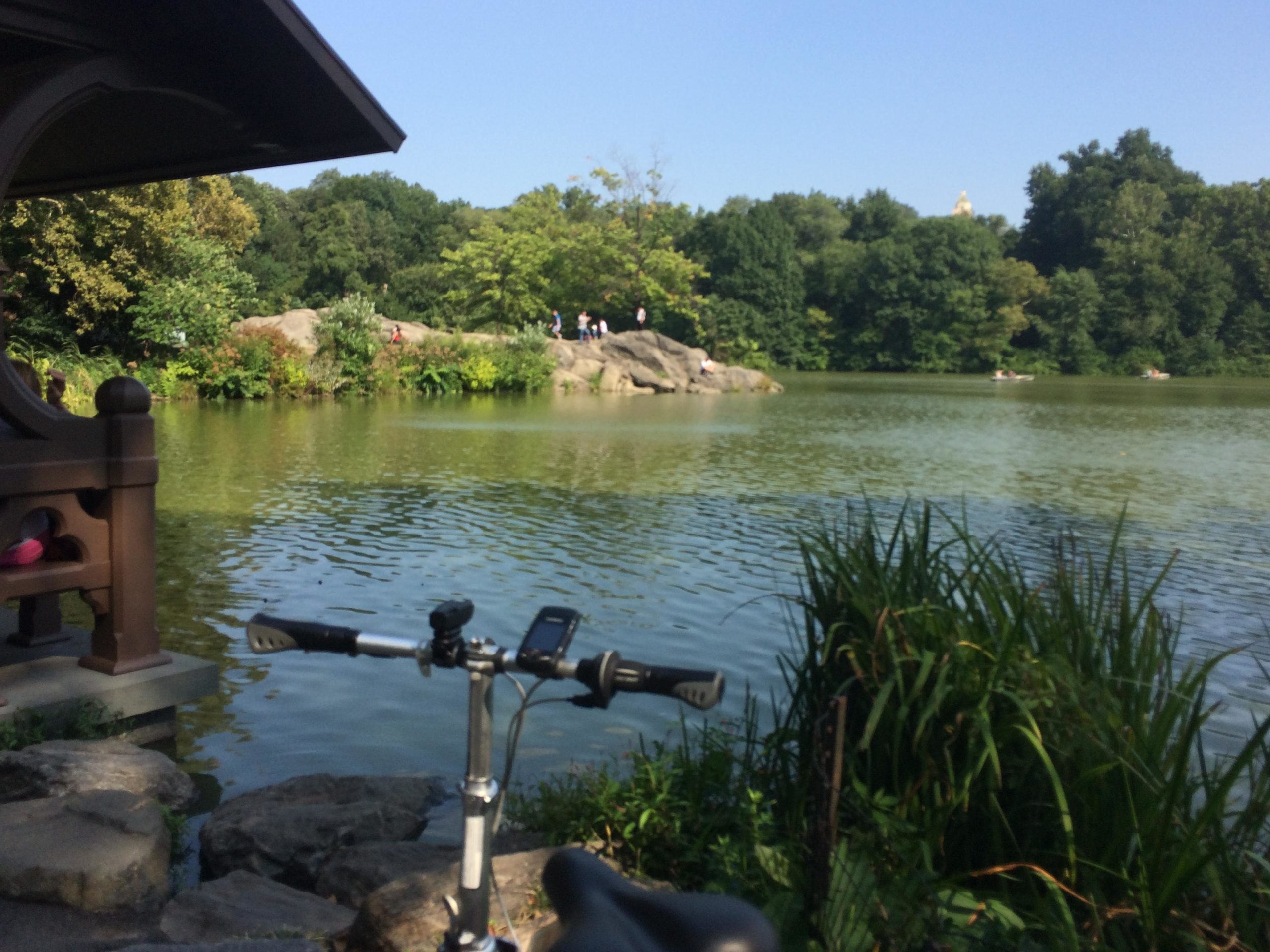 A serene view of a park lake surrounded by greenery, featuring a rock formation on the left and several people enjoying the outdoors in the distance. In the foreground, a bicycle rests on a rocky shore, adding to the peaceful atmosphere under a clear blue sky. Central Park Green way mountain bike trail.