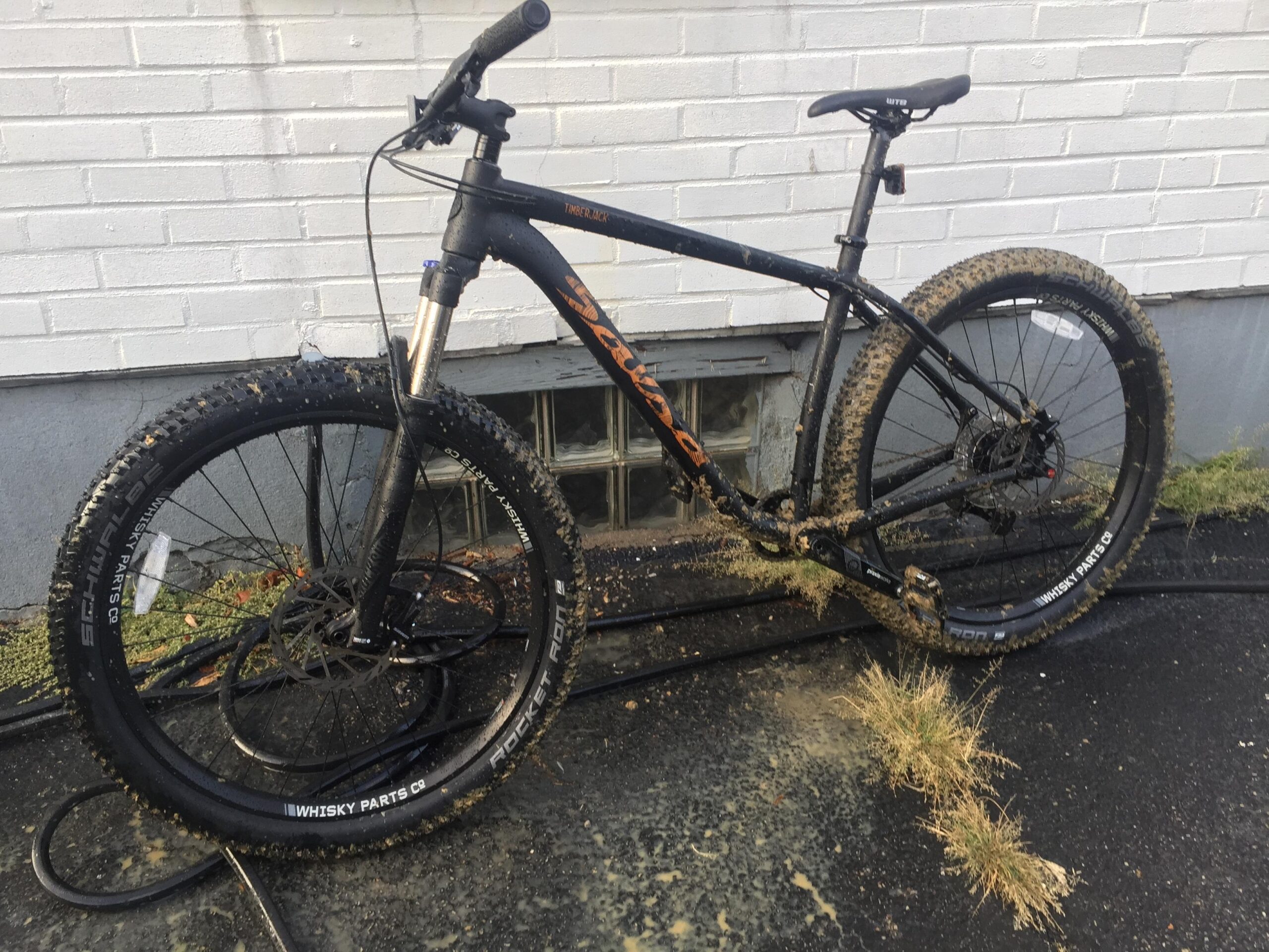 Salsa Timberjack: A mountain bike with a black frame, featuring thick tires covered in mud and dirt, is resting against a white brick wall. Some grass and dirt are splattered on the bike and the ground beneath it, indicating recent outdoor use.