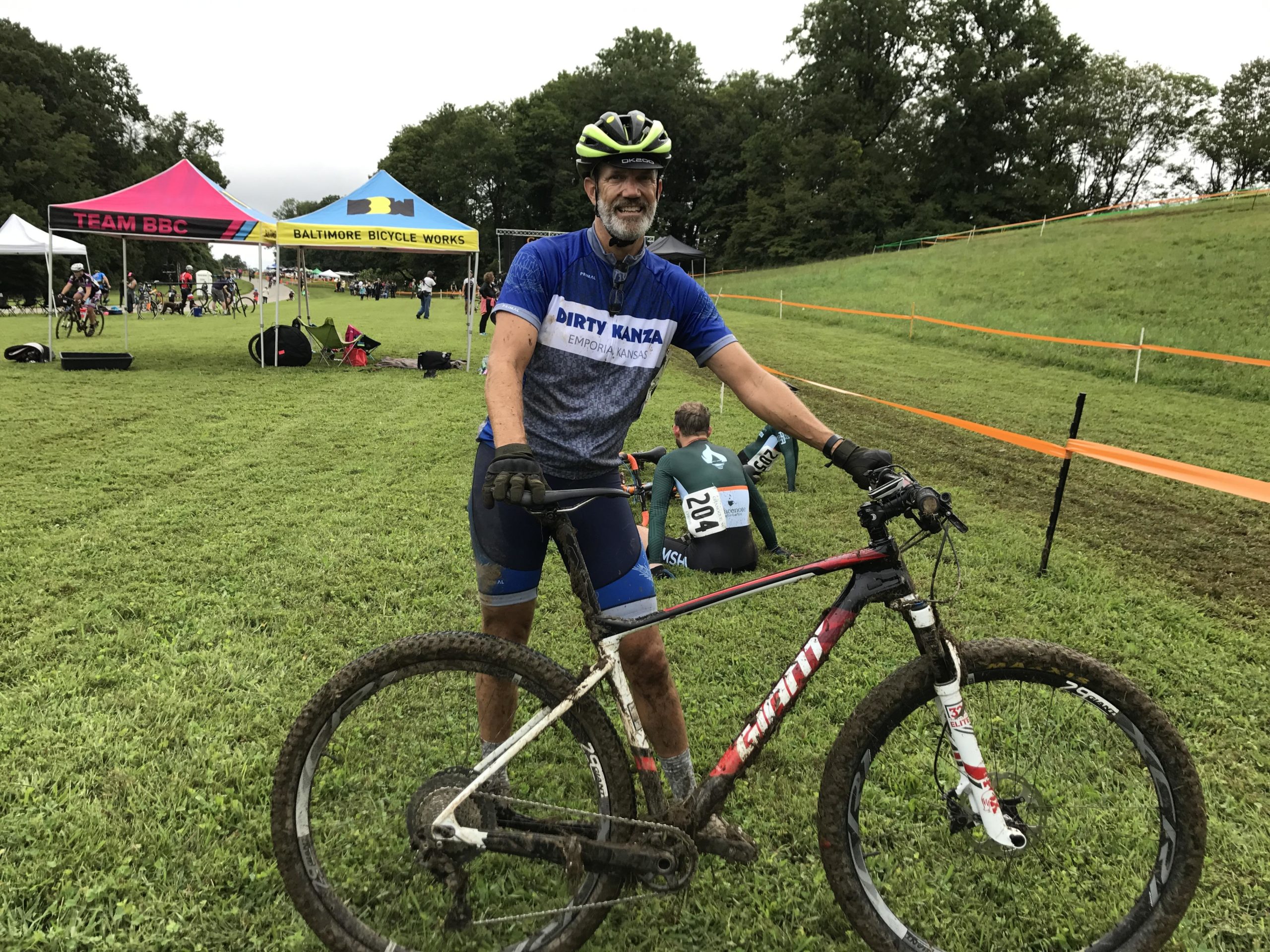 A smiling cyclist in a blue jersey with "Dirty Kanza" printed on it stands next to a muddy mountain bike. The background shows a grassy field with various tents for cycling teams, including Team BBC and Baltimore Bicycle Works, and other cyclists preparing for an event. The cyclist