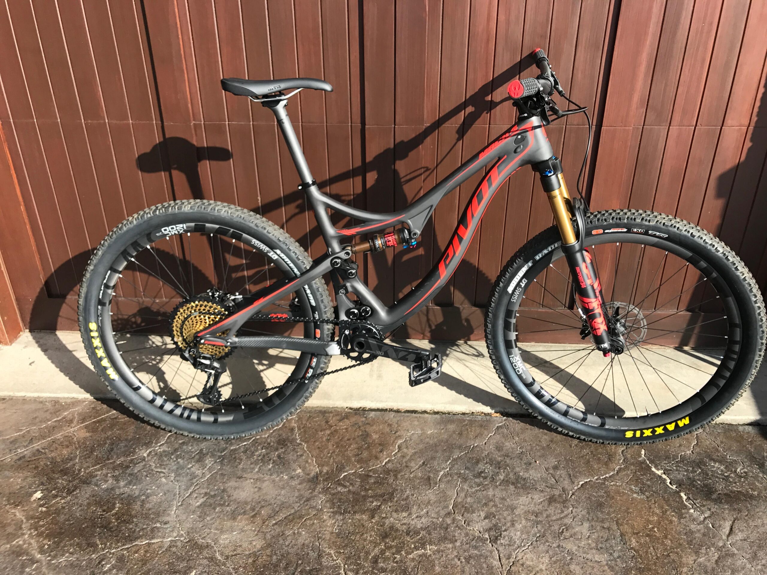 Pivot Mach 4: A sleek mountain bike with a gray frame and red accents, featuring large knobby tires and a gold rear cassette. The bike is positioned sideways against a wooden wall, showcasing its suspension fork and various components. The setting is well-lit, highlighting the bike's details and design.