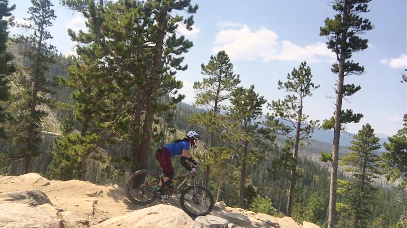A mountain biker navigating a rocky trail surrounded by tall pine trees and a clear blue sky. Trestle Bike Park mountain bike trail.
