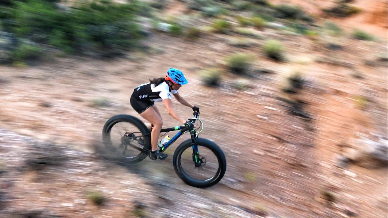 A cyclist riding a fat bike down a rocky trail, captured in motion with a blurred background to emphasize speed. The cyclist is wearing a blue helmet and a black and white cycling outfit. The terrain features dirt and sparse vegetation. Mariposa Fat Bike Trails mountain bike trail.