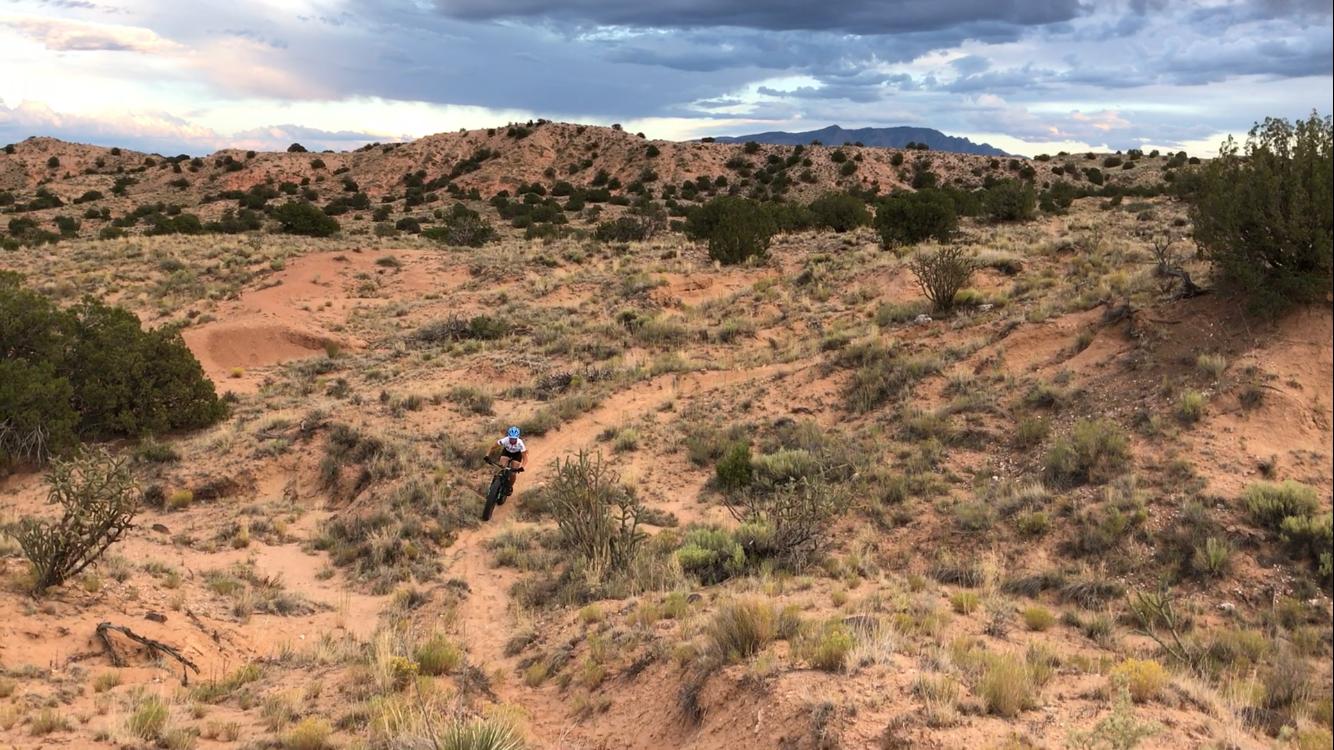 A mountain biker navigates a winding dirt trail surrounded by desert vegetation and rugged terrain, with hills and clouds in the background. Mariposa Fat Bike Trails mountain bike trail.