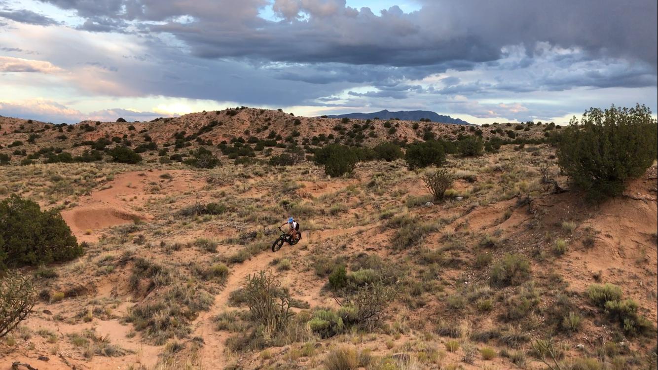 A mountain biker riding along a dirt trail in a hilly desert landscape, surrounded by sparse vegetation and under a cloudy sky. Mariposa Fat Bike Trails mountain bike trail.