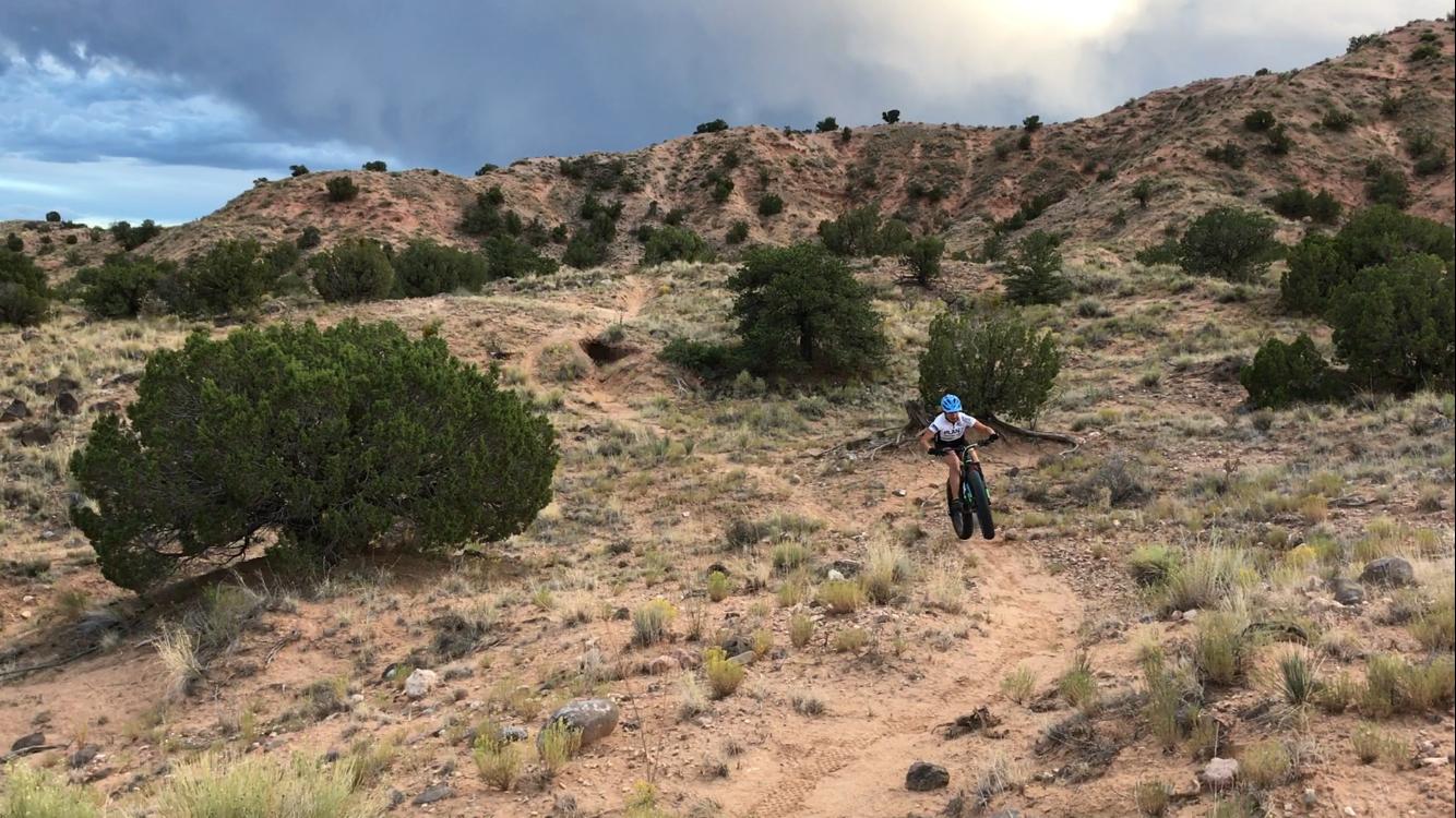 A person riding a mountain bike on a dirt trail through a rugged, arid landscape featuring shrubs and small hills under a cloudy sky. Mariposa Fat Bike Trails mountain bike trail.