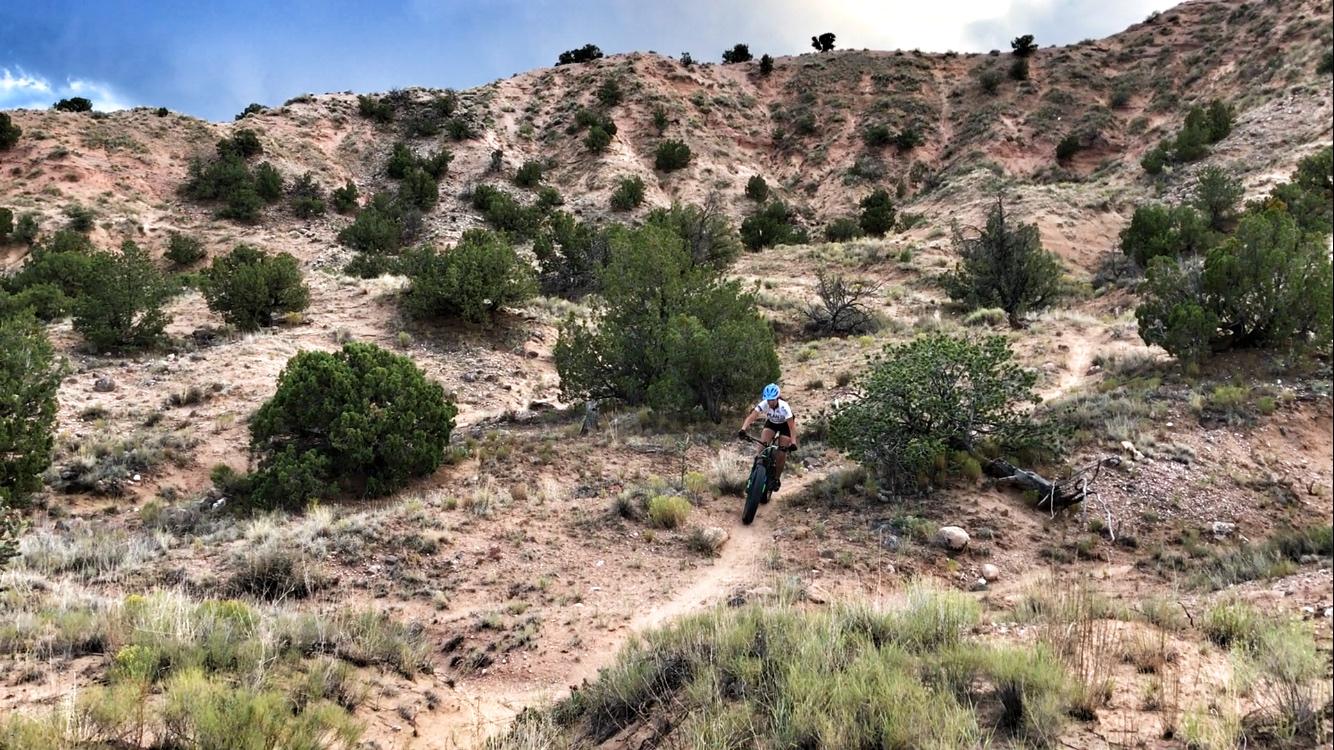 A mountain biker riding down a dirt trail surrounded by shrubs and rocky terrain, with a steep hill in the background under a partly cloudy sky. Mariposa Fat Bike Trails mountain bike trail.