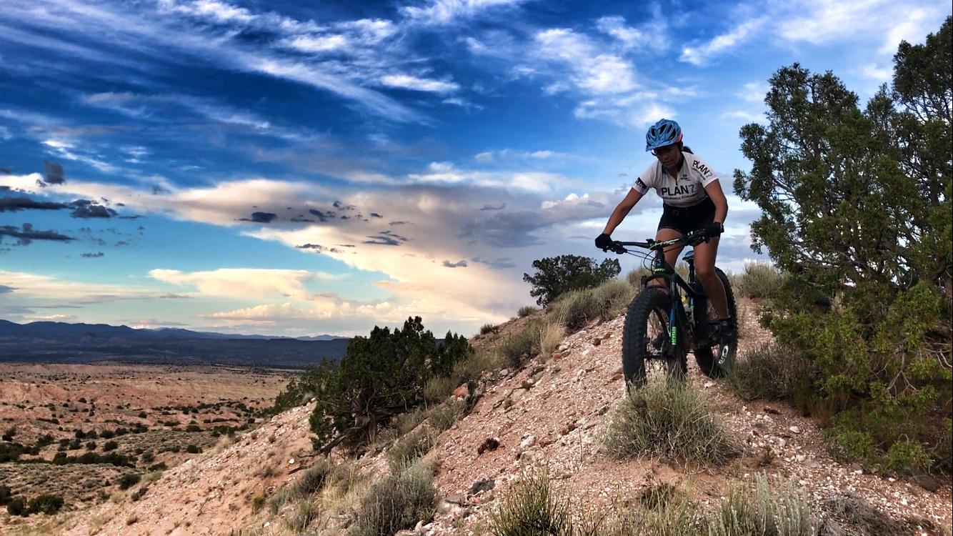 A mountain biker navigating a rocky trail in a desert landscape, with expansive skies filled with clouds in the background. The rider is wearing a helmet and a cycling jersey while focused on maintaining balance on the gravel path. Mariposa Fat Bike Trails mountain bike trail.