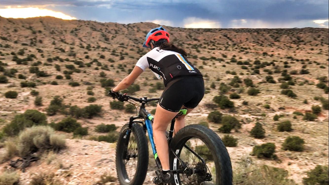 A person riding a mountain bike on a dirt trail surrounded by a desert landscape with low shrubs and rolling hills under an overcast sky. The cyclist is wearing a helmet and cycling outfit, focused on navigating the terrain. Mariposa Fat Bike Trails mountain bike trail.
