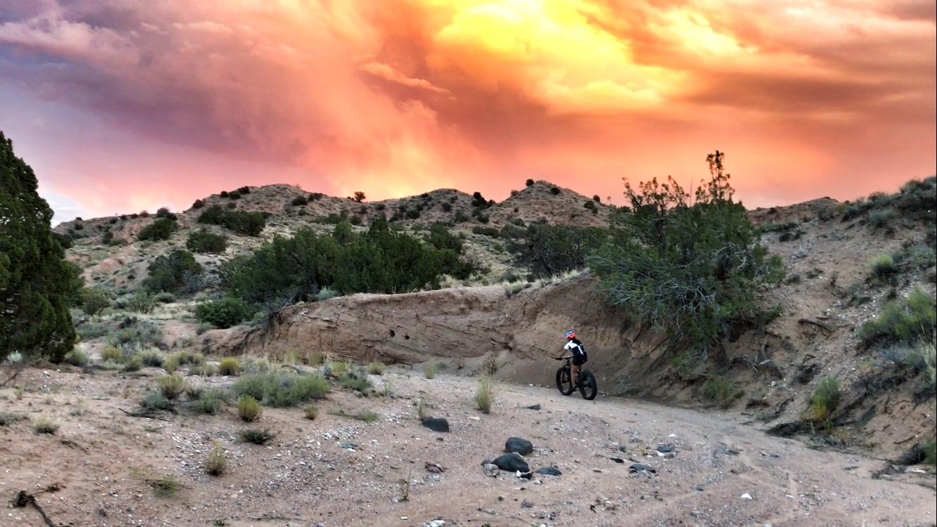 A mountain biker riding along a dirt trail in a desert landscape, with vibrant orange and pink clouds in the sky during sunset. Mariposa Fat Bike Trails mountain bike trail.