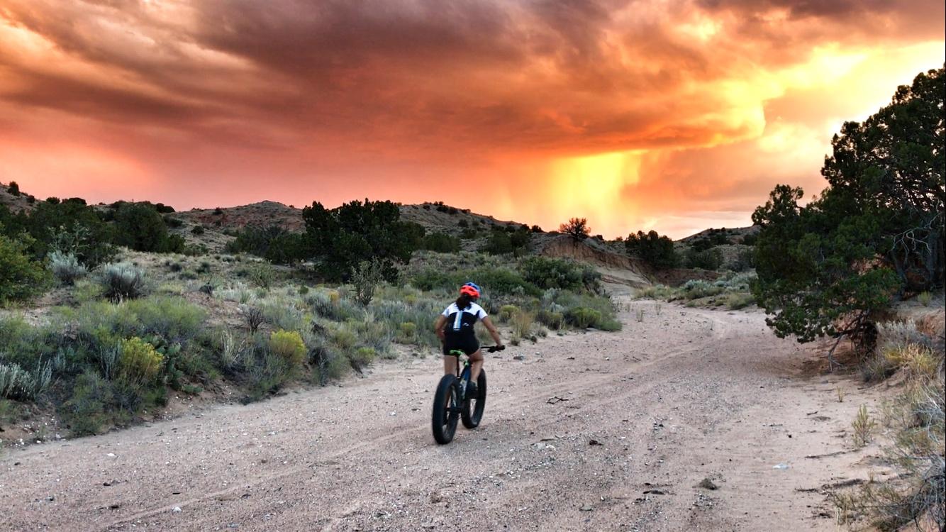 A person riding a fat bike on a sandy trail surrounded by shrubs and small trees, with a dramatic sunset sky filled with orange, pink, and purple clouds in the background. Mariposa Fat Bike Trails mountain bike trail.