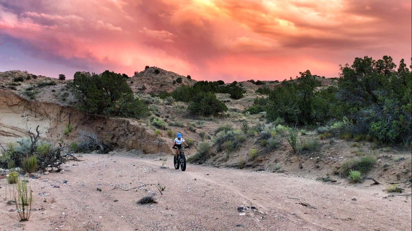 A mountain biker rides along a sandy trail surrounded by desert vegetation, with hills and a vibrant sunset sky in shades of pink and orange in the background. Mariposa Fat Bike Trails mountain bike trail.