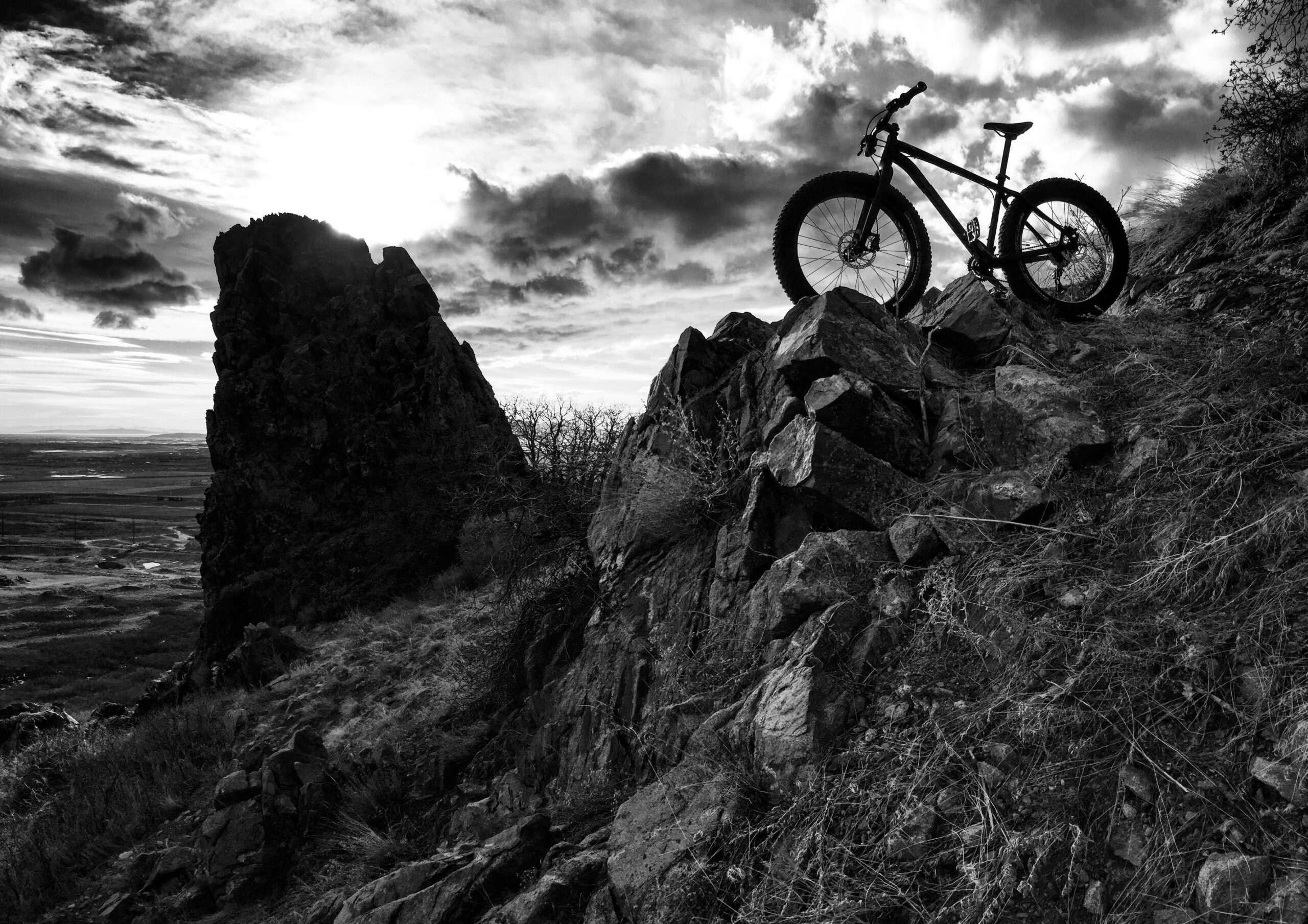 Specialized Fatboy: A black and white image of a mountain bike positioned on a rocky outcrop, silhouetted against a dramatic sky with clouds. The sun is setting in the background, casting a bright light around the bike and highlighting the rugged terrain.