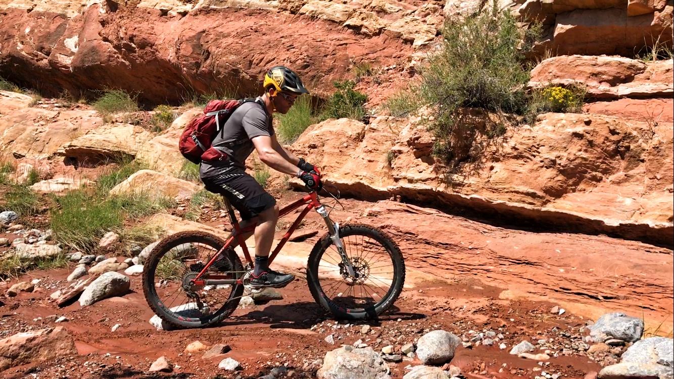 A mountain biker navigating a rocky trail with red rock formations in the background. The rider is wearing a helmet and gloves, with a backpack, and is positioned on a bicycle as they traverse a dirt path dotted with stones and vegetation. Golden Open Space mountain bike trail.