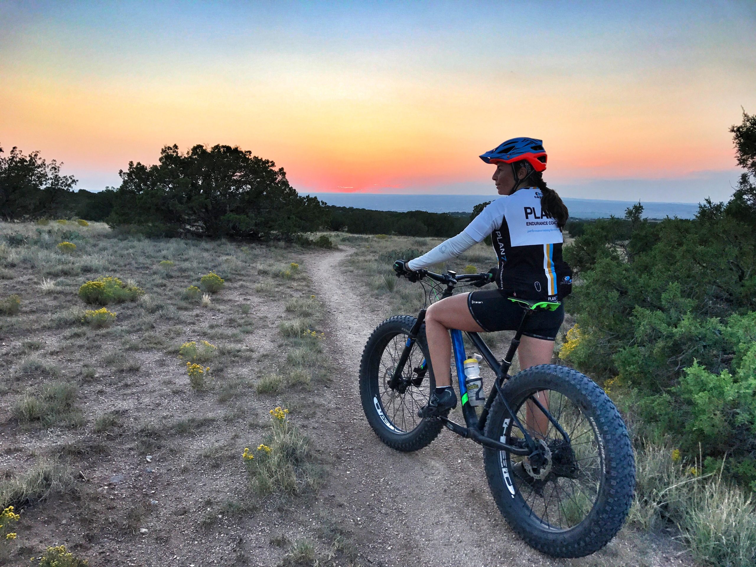 A cyclist wearing a helmet and cycling gear stops on a gravel trail surrounded by a grassy landscape and sparse shrubs, with a vibrant sunset in the background that features hues of orange and blue. Placitas Trails mountain bike trail.