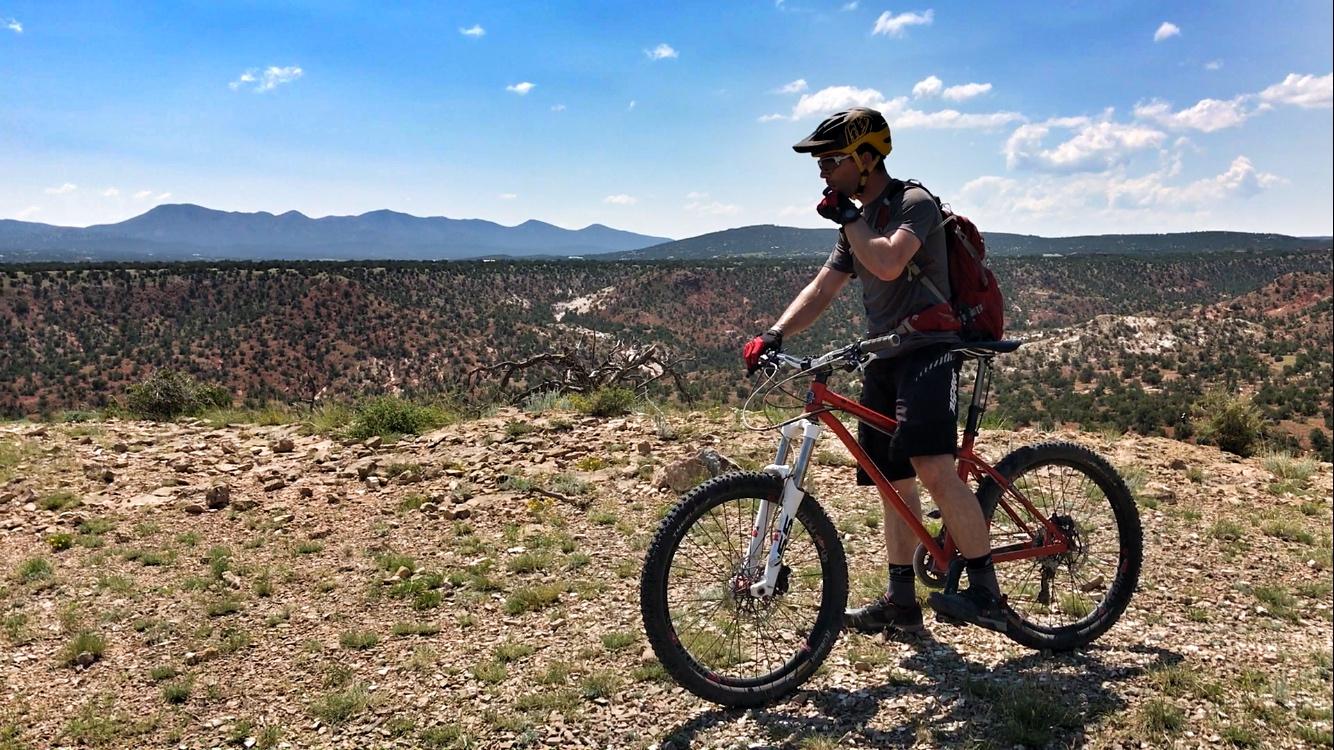 A mountain biker wearing a helmet and gloves stands next to a red bicycle, overlooking a scenic landscape of rolling hills and mountains under a partly cloudy sky. The terrain features rocky ground and sparse vegetation, highlighting a natural environment. Golden Open Space mountain bike trail.
