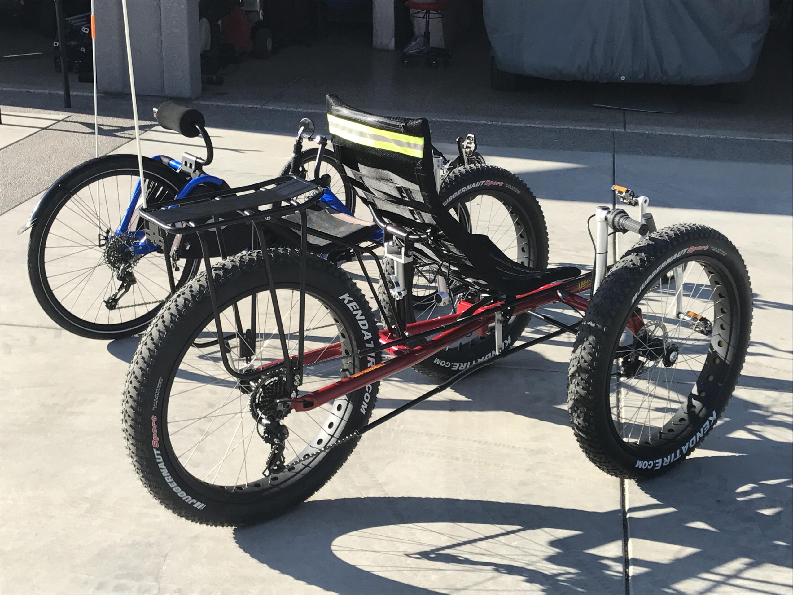 Santa Cruz Bronson: A red three-wheeled bicycle with large, knobby tires is positioned prominently in the foreground, featuring a reclining seat and a rear storage rack. Behind it, a blue bicycle is partially visible, highlighting its traditional two-wheeled design. The setting appears to be a garage or driveway with a concrete surface.