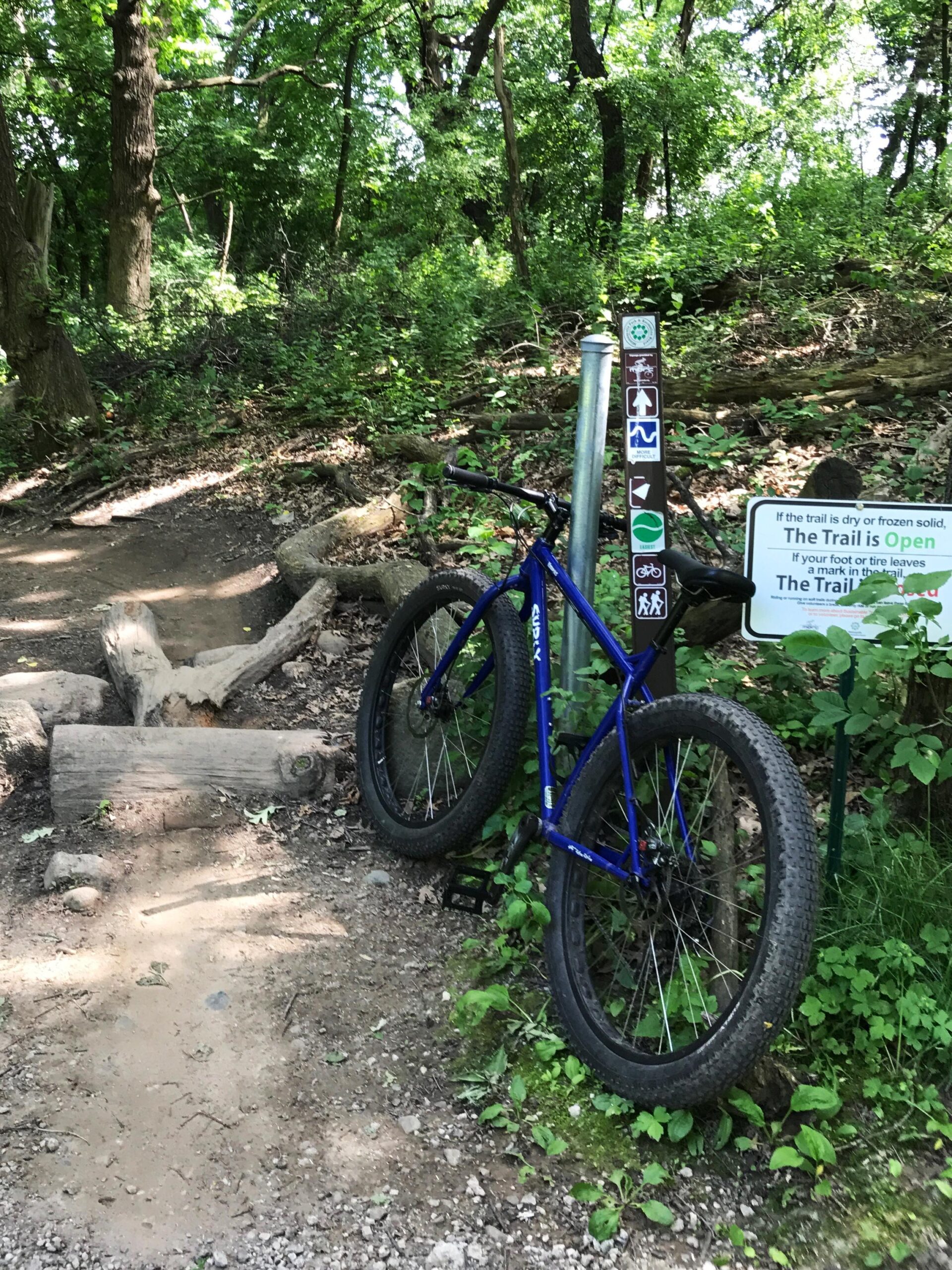 Surly Krampus: A blue mountain bike resting against a post near a forest trail. The trail is surrounded by green foliage, and there is a sign indicating trail rules and conditions. The path has dirt and small rocks, with some logs and natural features visible.