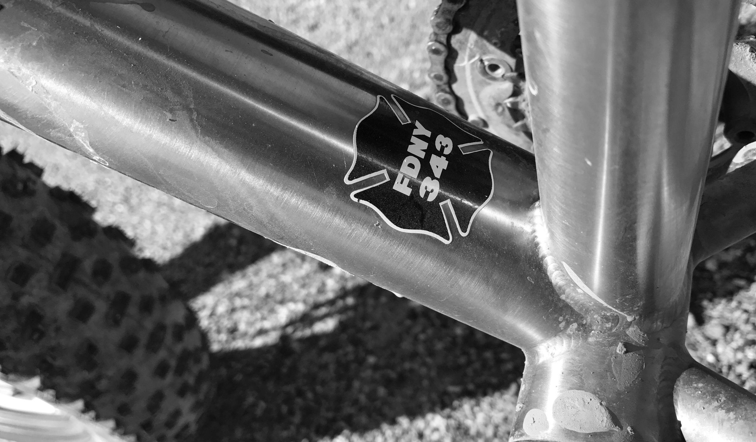 Close-up of a metallic bicycle frame featuring a sticker with the letters "FDNY" and the number "343," set against a blurred background of a bicycle tire. The image is in black and white, highlighting the frame's surface texture and the sticker's design. Mariposa Fat Bike Trails mountain bike trail.