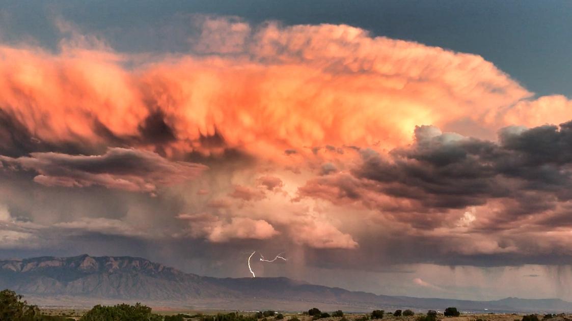 Dramatic clouds illuminated by a sunset with shades of orange and pink, looming over distant mountains. A flash of lightning is visible in the center of the image, while rain can be seen falling in the lower section. The scene captures the beauty and intensity of a stormy sky. Parkway Fatbike trail mountain bike trail.