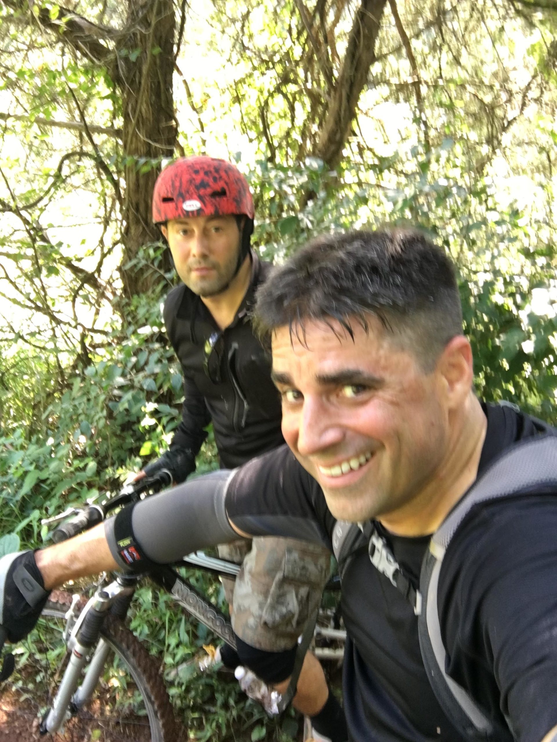 Two men taking a selfie while mountain biking on a wooded trail. One is wearing a red helmet with a black shirt and the other is dressed in a black long-sleeve shirt. They are surrounded by greenery and appear to be enjoying their outdoor activity. Six Mile Run mountain bike trail.