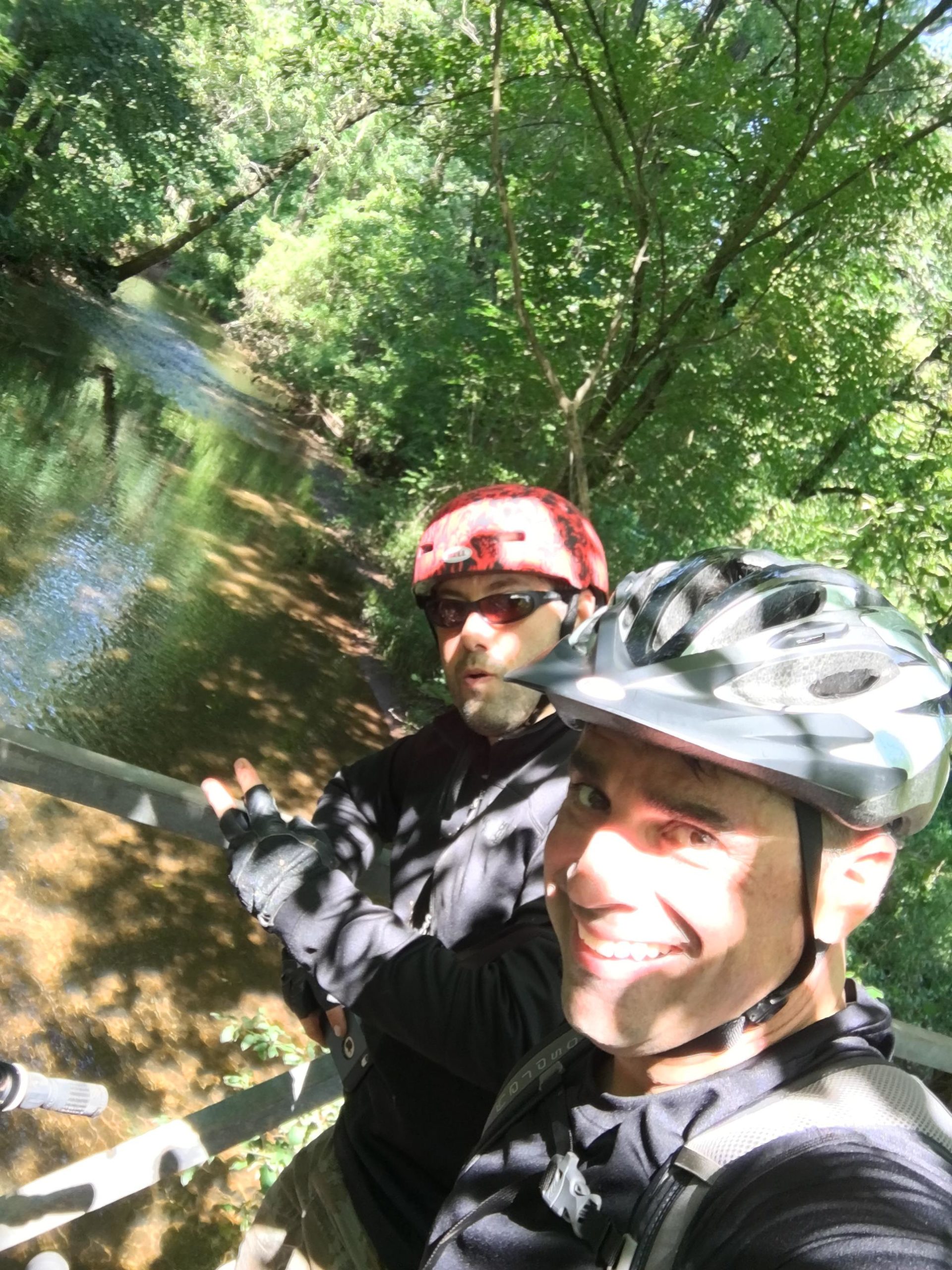 Two cyclists with helmets stand on a bridge over a stream, surrounded by lush greenery. One appears to be smiling and posing for the camera, while the other makes a playful expression. Bright sunlight filters through the trees, casting dappled shadows on the scene. Six Mile Run mountain bike trail.