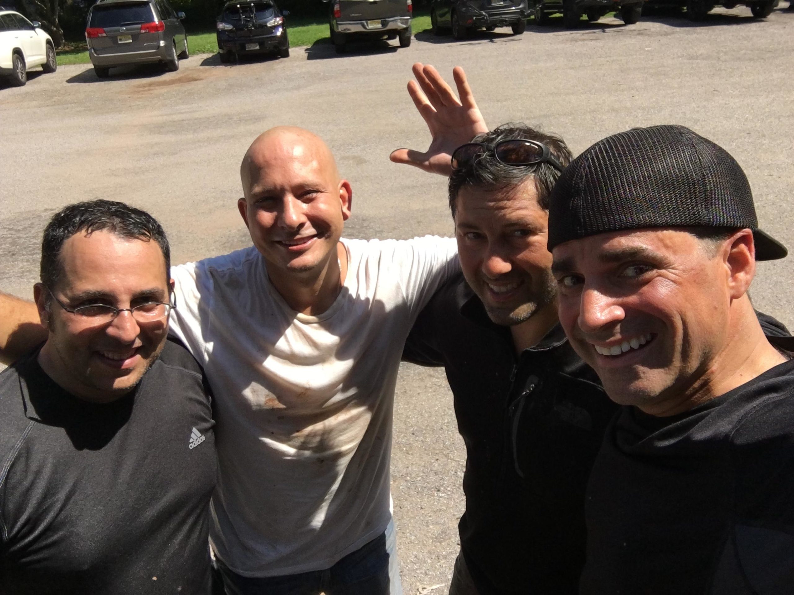 A group of four men standing together, smiling and posing for the camera in a sunny outdoor setting. They are wearing casual clothing, some with visible dirt or stains, suggesting they have been engaged in an outdoor activity. In the background, several parked vehicles can be seen on a gravel surface. Six Mile Run mountain bike trail.