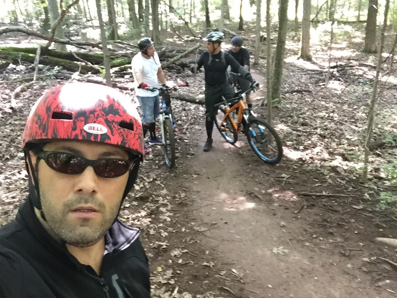 Three mountain bikers take a break on a wooded trail. One person in the foreground is wearing a red helmet and sunglasses, while two others are standing with their bikes in the background. The trail is surrounded by trees and scattered leaves, indicating a lush, natural environment. Six Mile Run mountain bike trail.