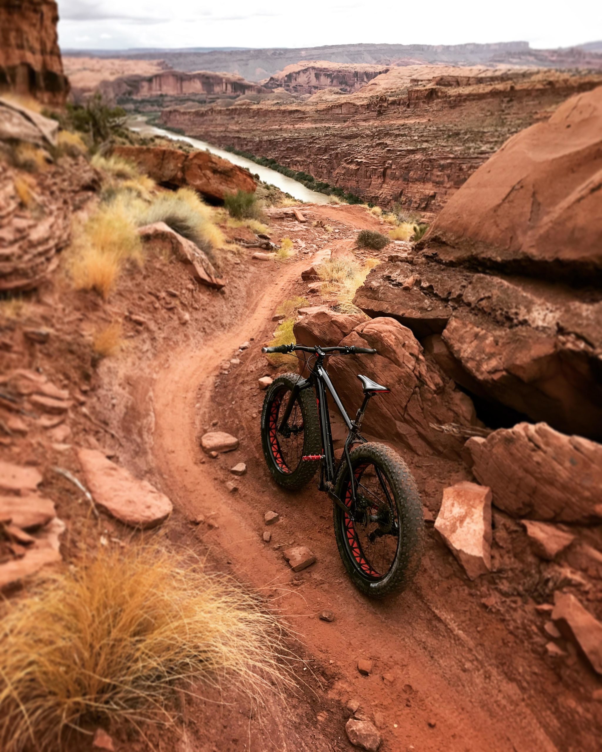 A black fat bike with red accents rests on a narrow dirt trail winding through rocky terrain, overlooking a river below and surrounded by dramatic red rock formations under an overcast sky. The Whole Enchilada mountain bike trail.
