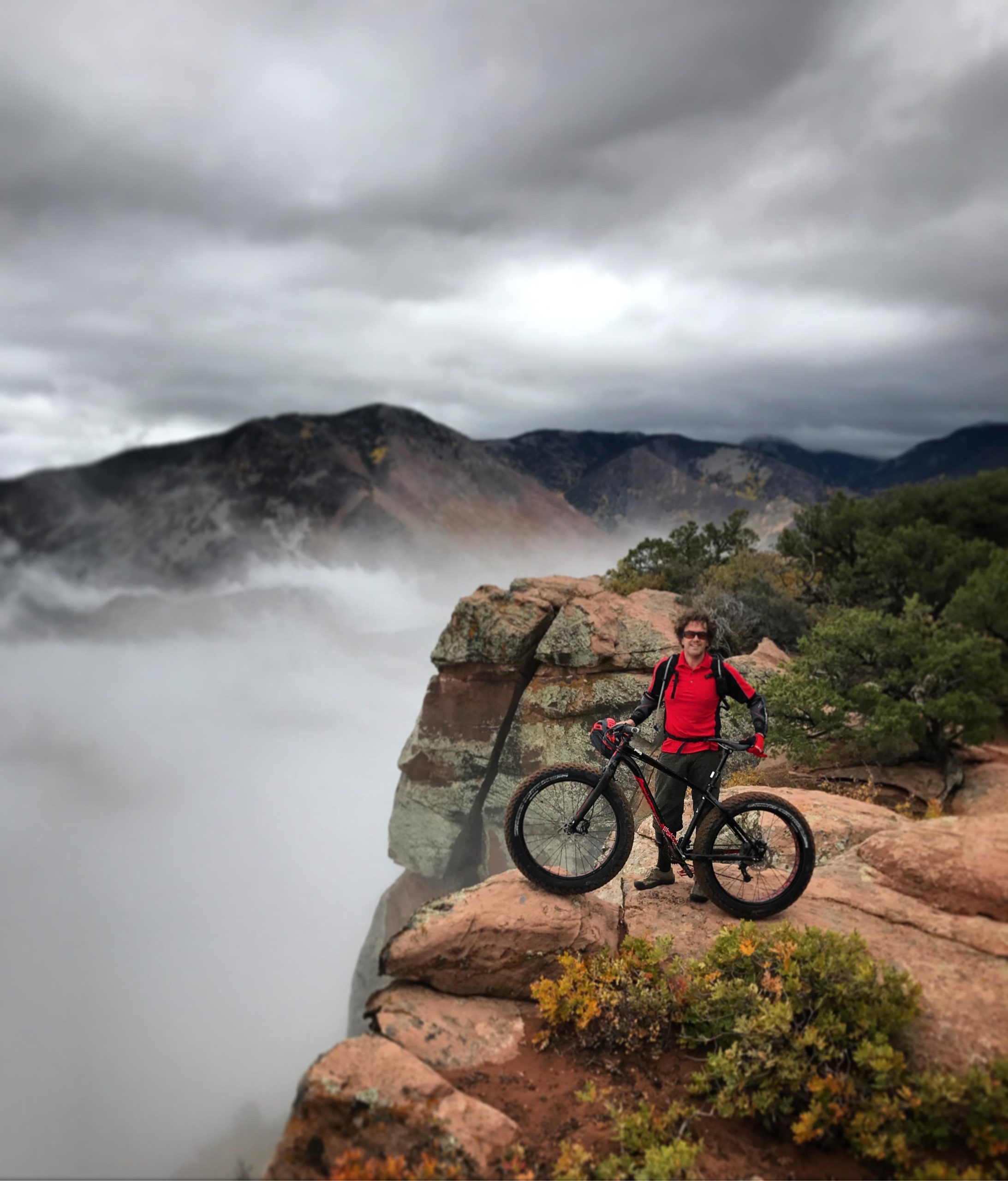 A person in a red shirt and black jacket stands proudly on a rocky outcrop, holding a black mountain bike. The background features dramatic mountains partially obscured by fog, with gray clouds overhead and hints of greenery on the rocky terrain. Porcupine Rim mountain bike trail.