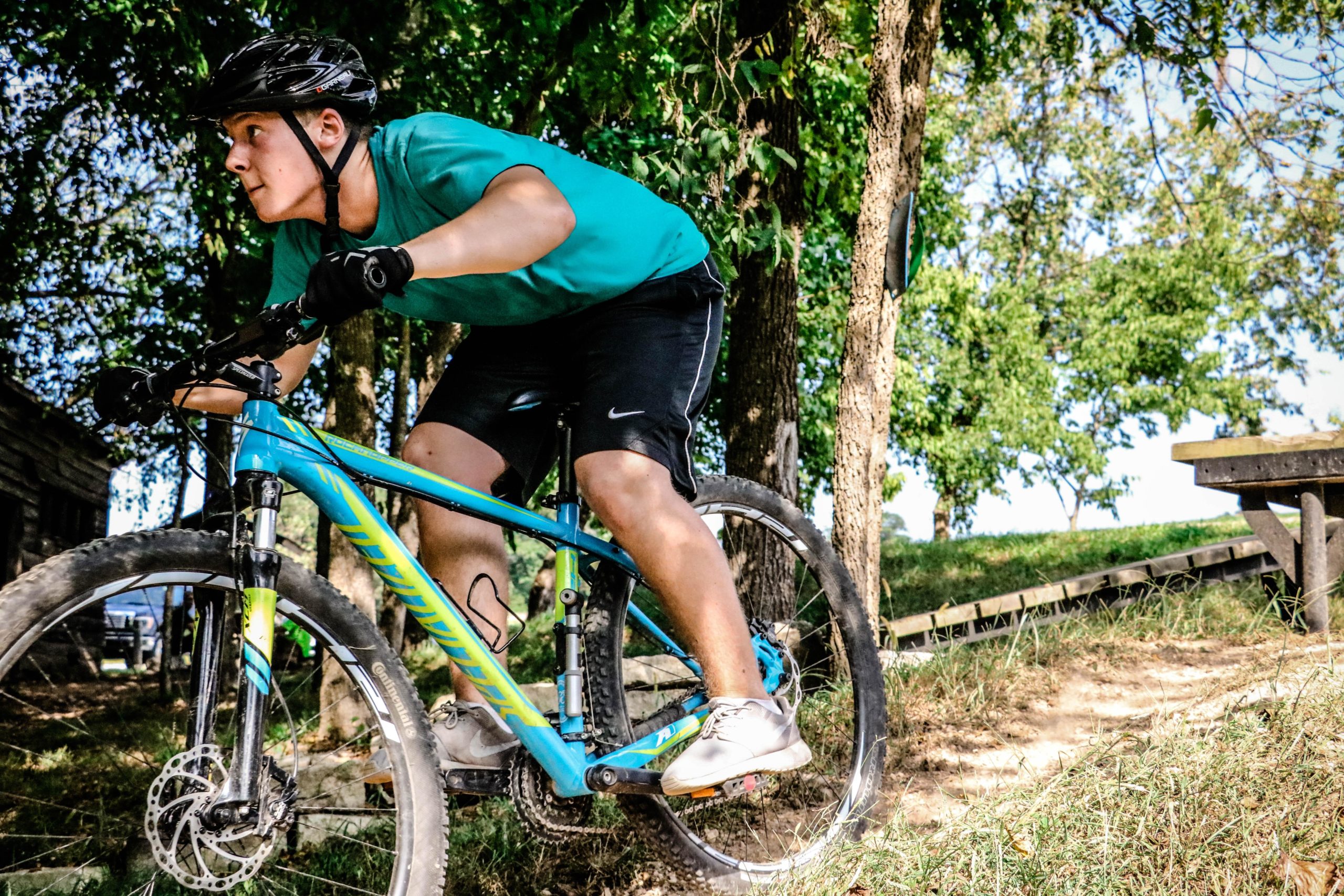 A young person in a teal shirt and black shorts riding a blue mountain bike, leaning forward as they navigate a grassy trail surrounded by trees. The rider is focused, wearing a helmet and gloves, with dirt and foliage visible in the background. Two Rivers Bike Park mountain bike trail.