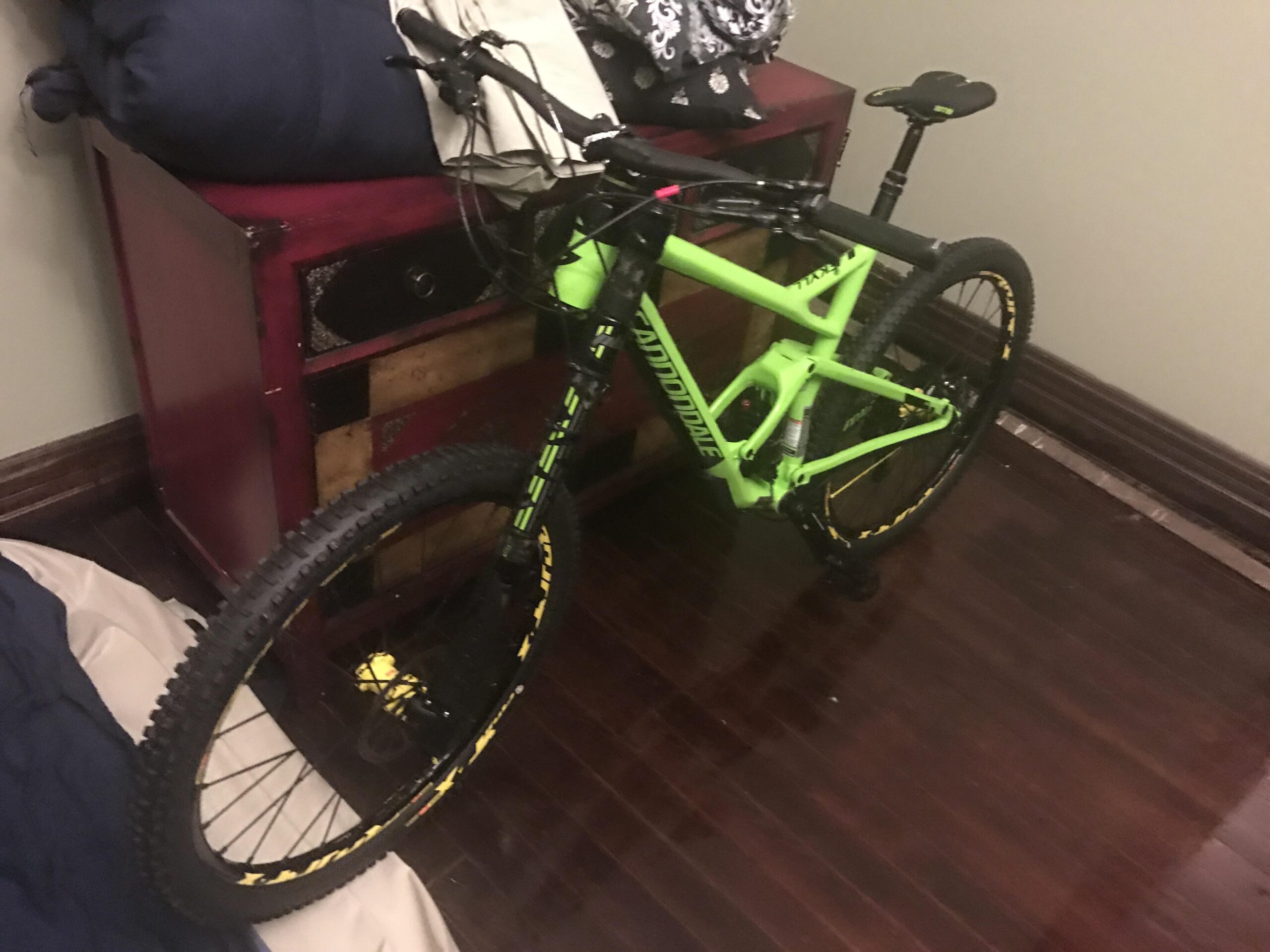 Cannondale Jekyll Carbon 1: A bright green mountain bike leans against a wooden chest in a room with dark hardwood flooring. The bike features thick, textured tires with yellow accents and is positioned near a pile of cushions. The background shows a light-colored wall, contributing to the casual indoor setting.