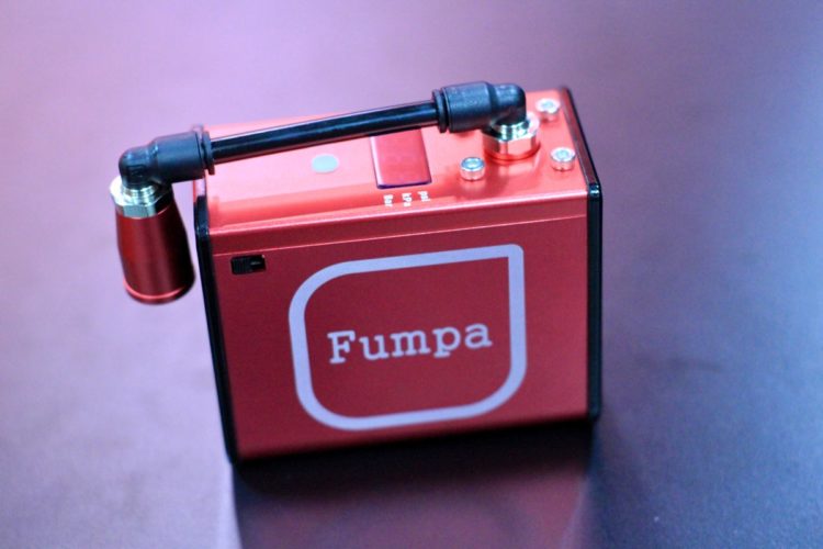 A small, red electronic device labeled "Fumpa" on its front, featuring various knobs and a nozzle attached to a black tube. The device is placed on a dark surface, reflecting light.