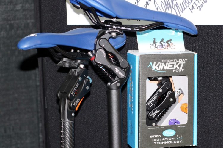 A blue bicycle saddle mounted on a Kinekt post with visible suspension features, alongside a box labeled "BODYFLOAT KINEKT POST," showcasing its body isolation technology. The background is black with a handwritten note partially visible.