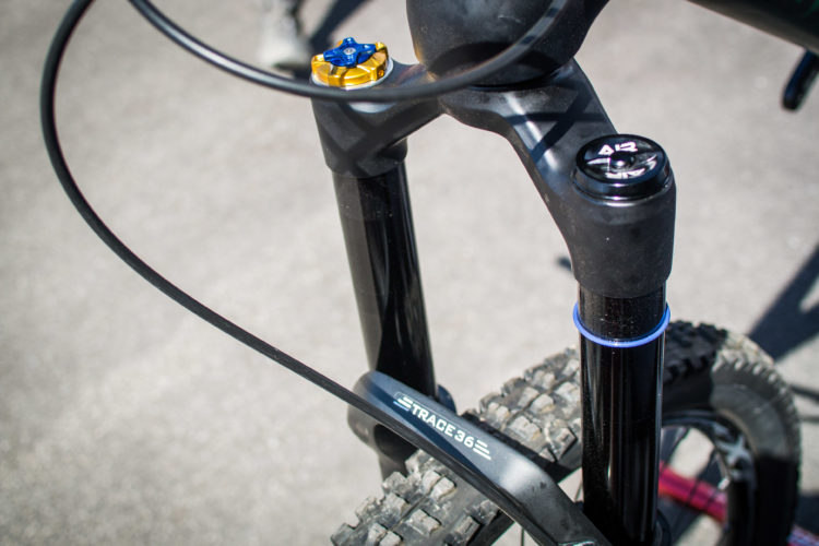 Close-up view of a mountain bike's front suspension fork, showcasing the components including adjustment knobs and a tire with knobby tread. The image highlights a logo labeled "TRACE 36" on the fork brace, set against a blurred background of pavement.
