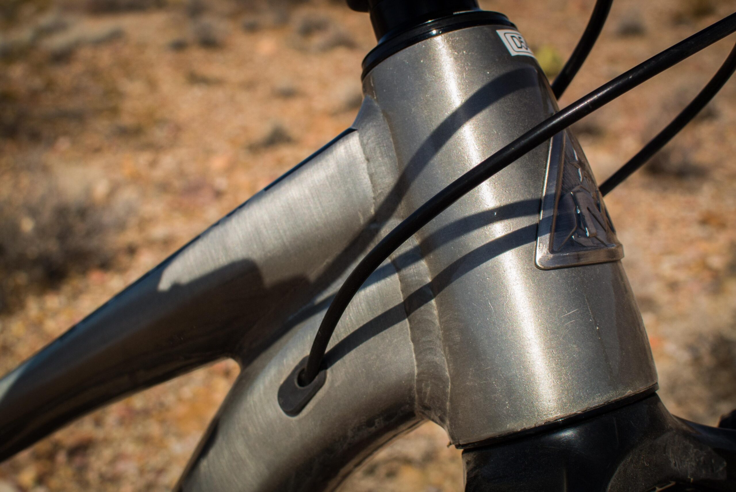 Marin B-17 3: Close-up view of a metal bicycle frame at the head tube, showcasing the welds and cable housing against a blurred natural background. The frame has a brushed finish with visible texture and sheen.