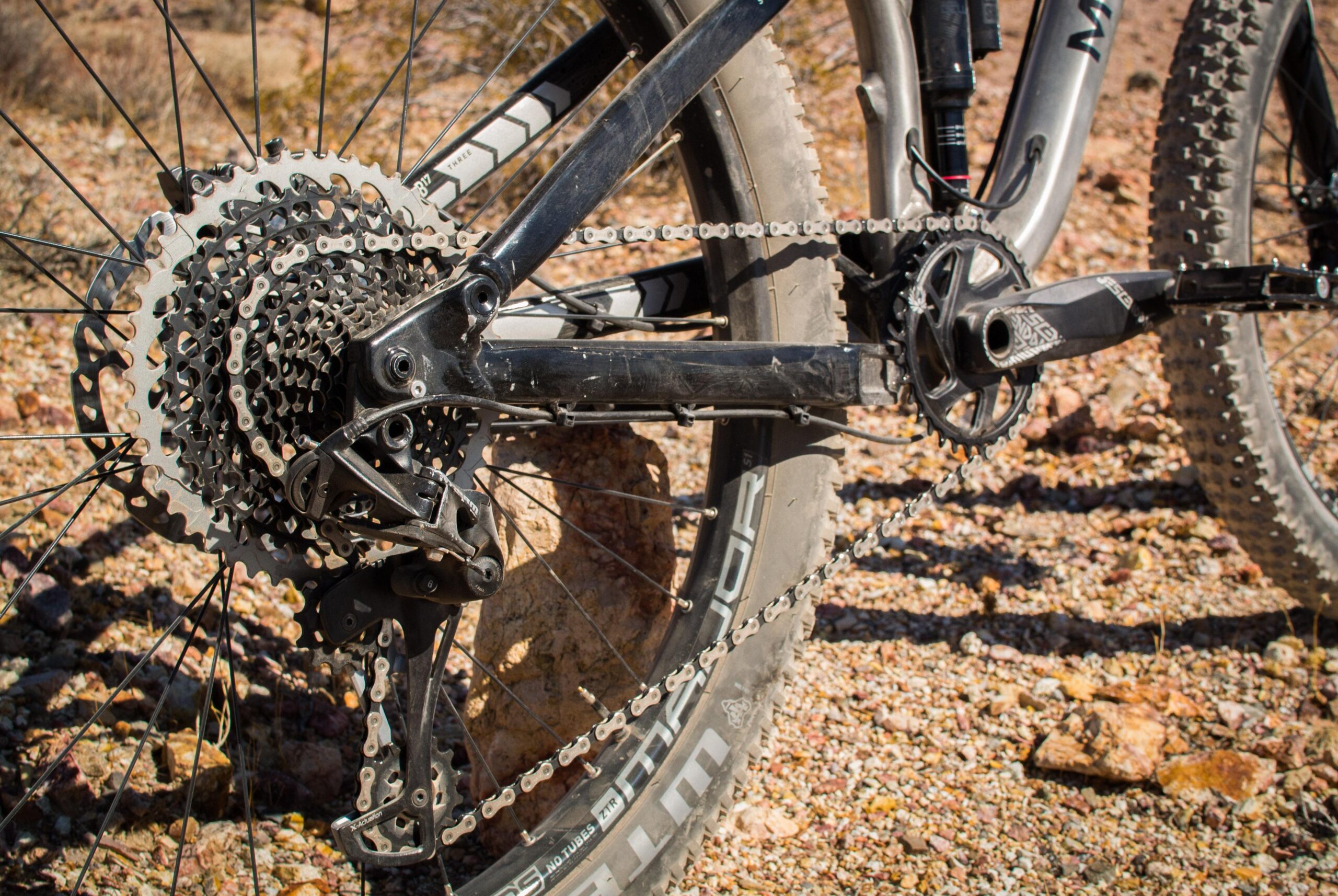 Marin B-17 3: An up-close view of a mountain bike's drivetrain, showcasing the cassette, chain, derailleur, and crankset, set against a rocky terrain. The bike's rear wheel is partially visible, highlighting the tire tread. The image captures the mechanical details of the bike, emphasizing its rugged design and components.