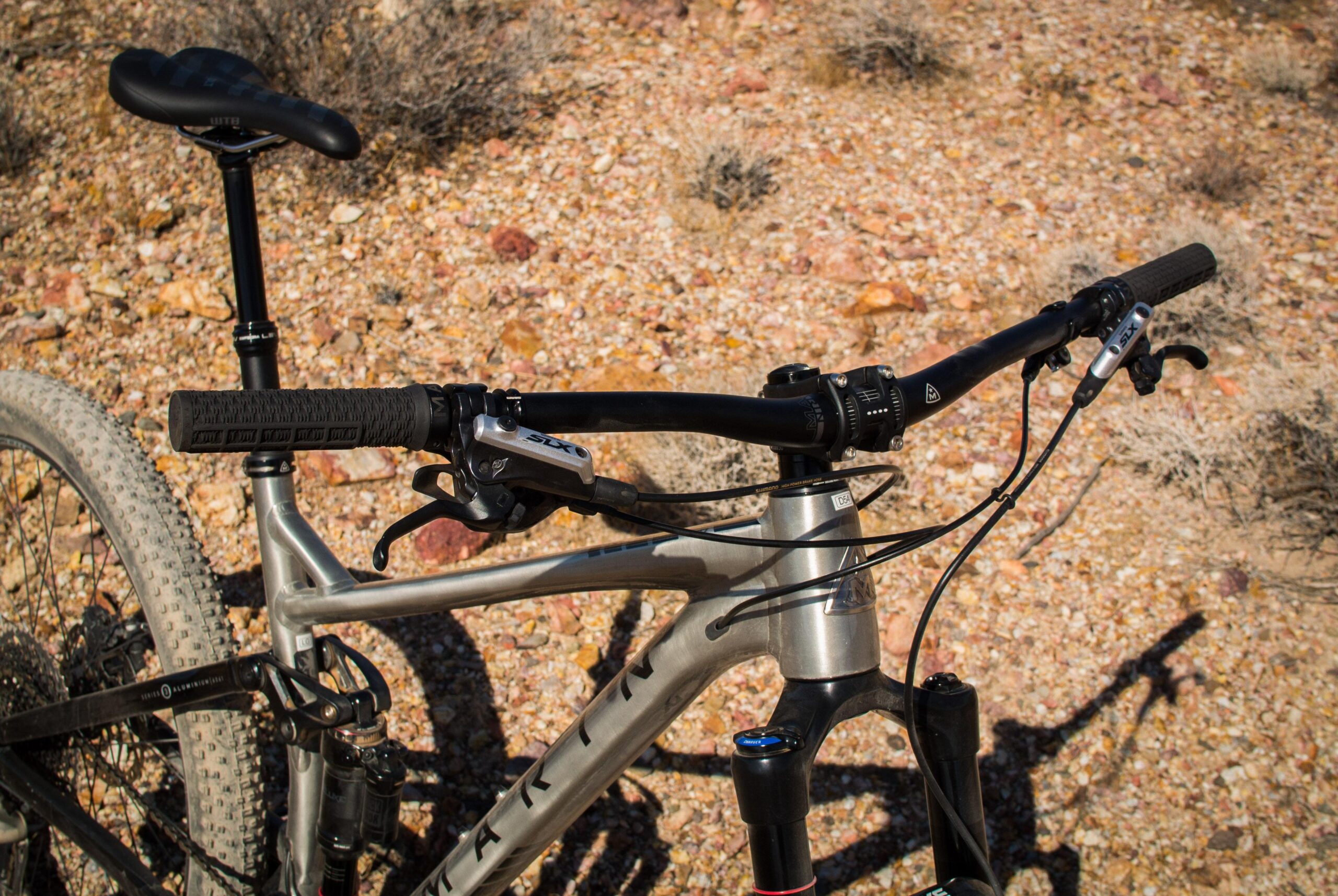 Marin B-17 3: Mountain bike handlebars and seat close-up, set against a rocky terrain backdrop. The bike features black grips, a silver brake lever, and a shiny metallic frame.